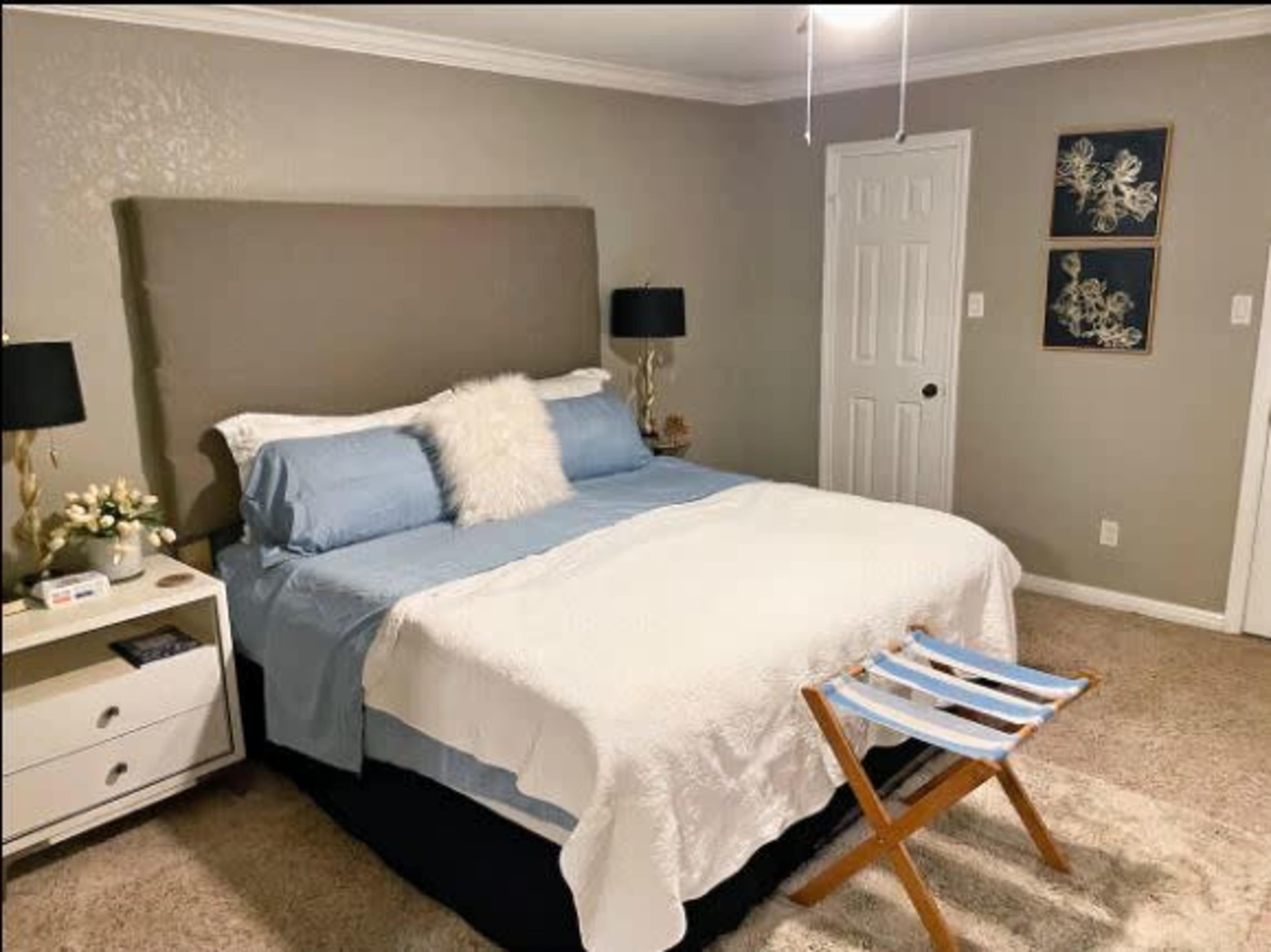 A neatly arranged bedroom features a bed with blue and white bedding, flanked by black lamps, a nightstand with decorative items, and a folding chair.
