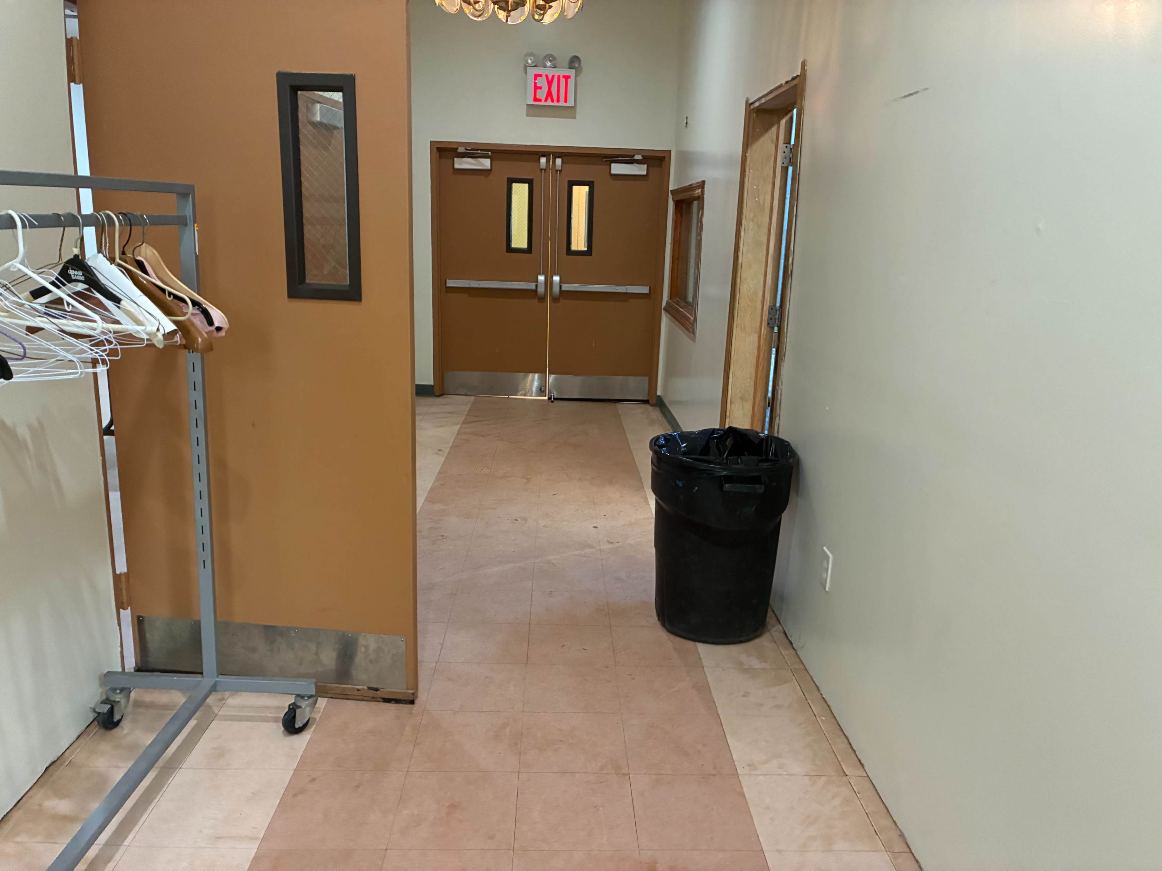 The image shows a hallway with a clothing rack on the left and double doors at the end, alongside a trash can on the floor.