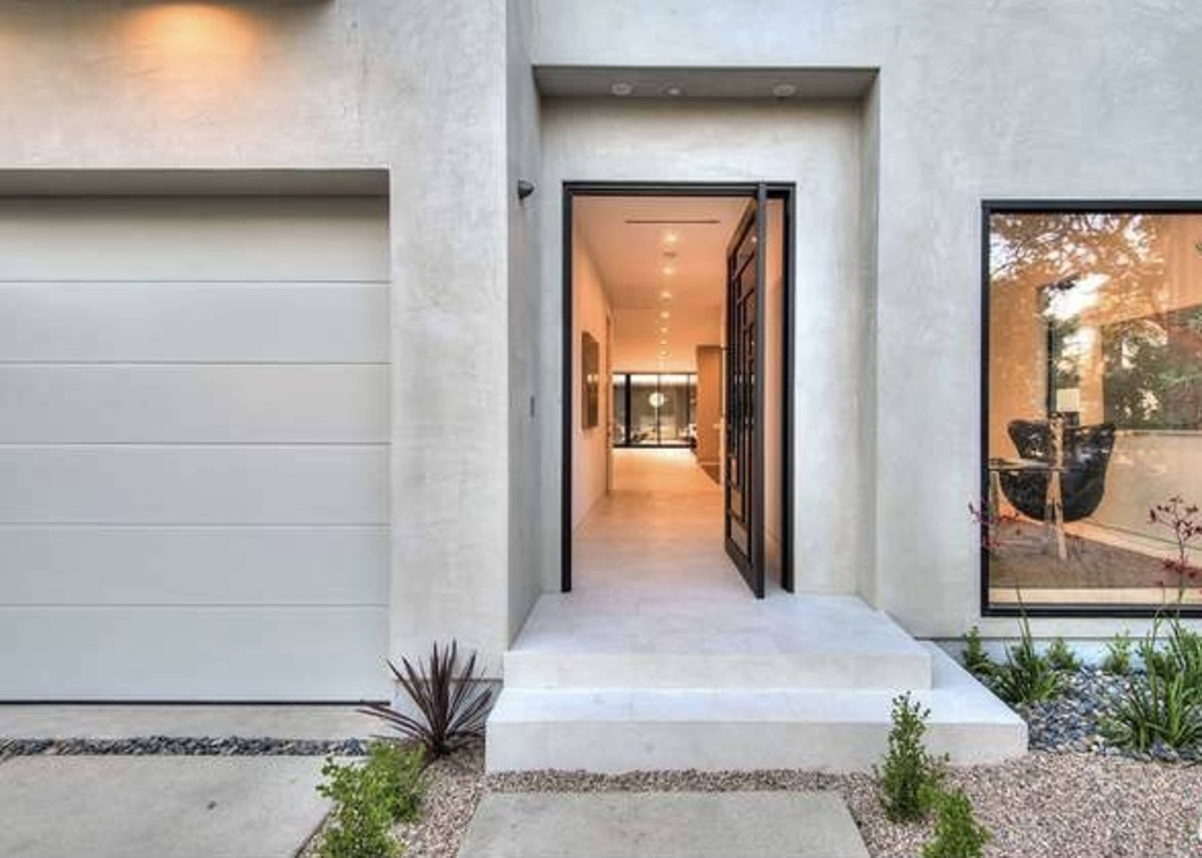 The image shows the entrance of a modern house with a large front door opening into a bright hallway, flanked by a garage and large windows.