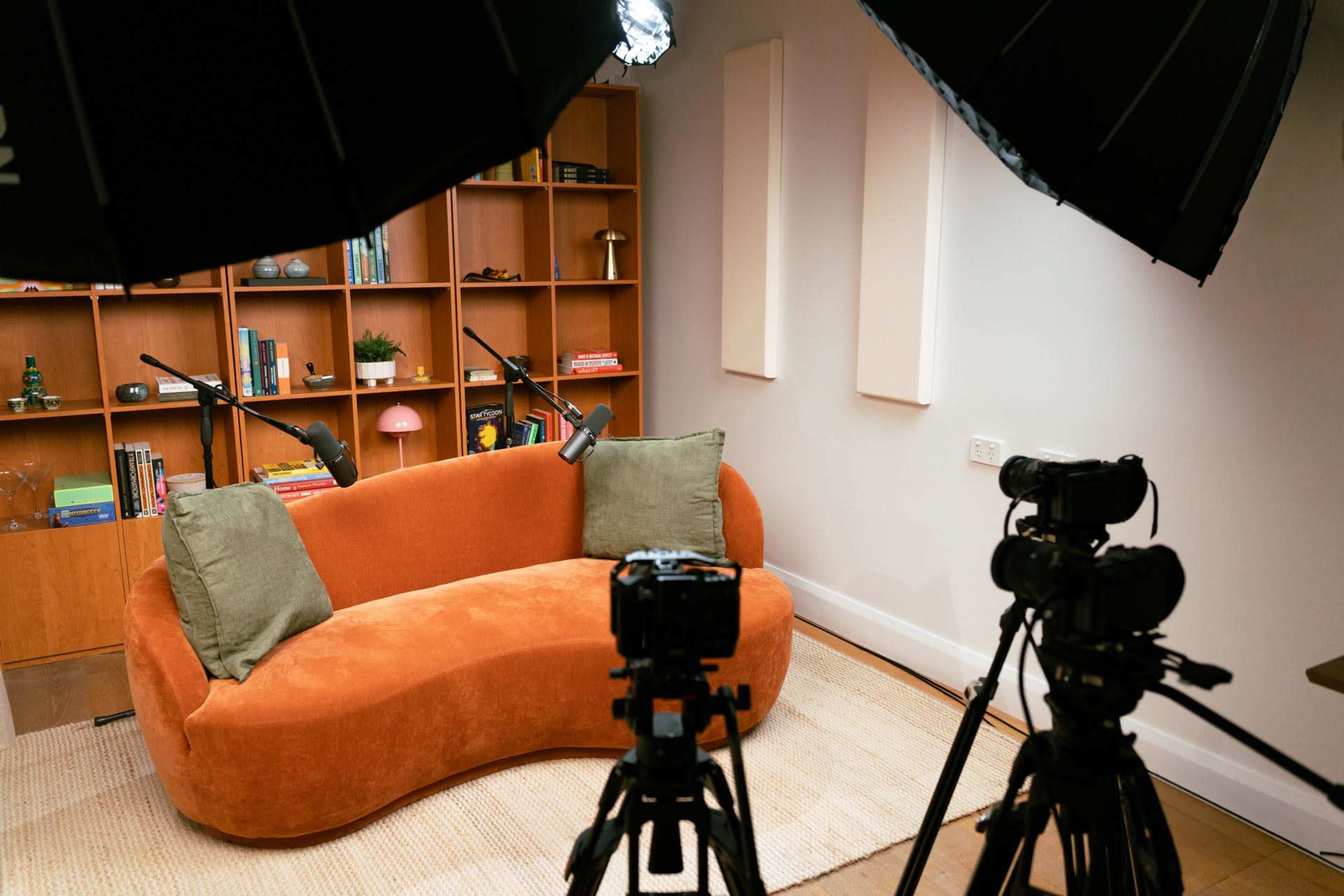 A cozy orange sofa with two green pillows is positioned in a well-lit room that features bookshelves and multiple cameras on tripods.