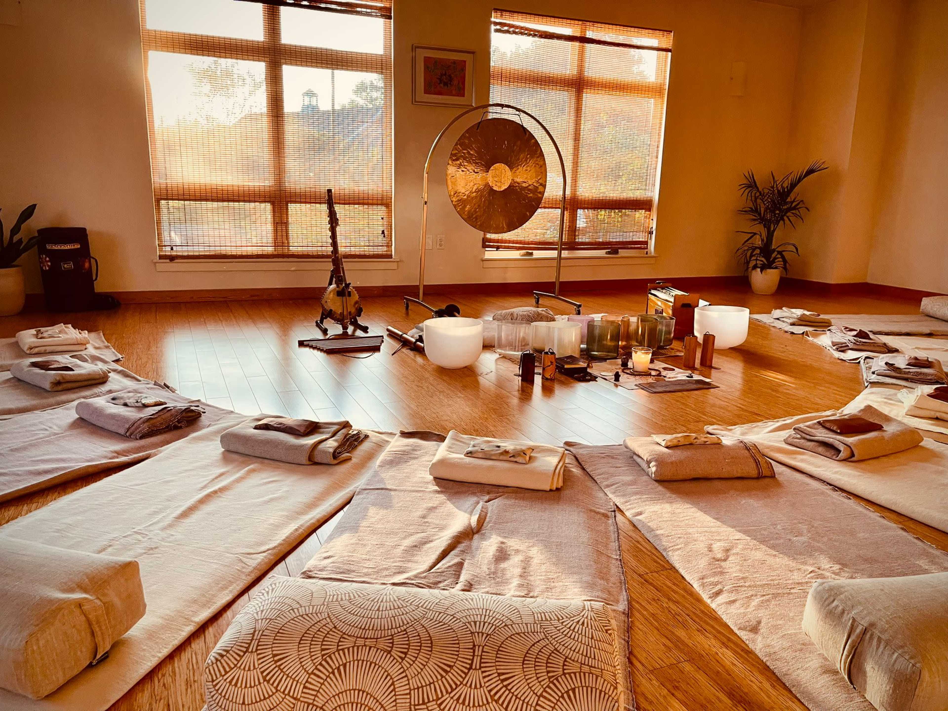 A serene yoga studio is set up with evenly arranged mats, cushions, and sound healing instruments in the center, illuminated by warm morning light filtering through large windows.
