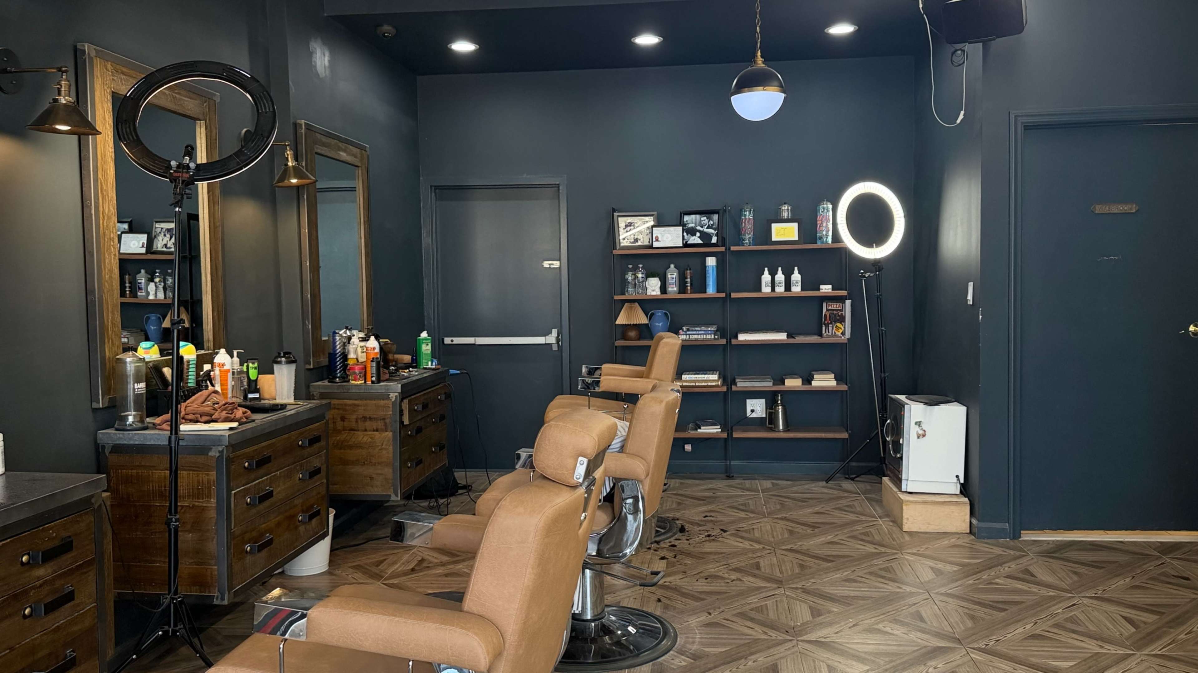 The image shows a barber shop with two vintage-style chairs, a lighted mirror, shelves displaying grooming products, and a dark blue wall.