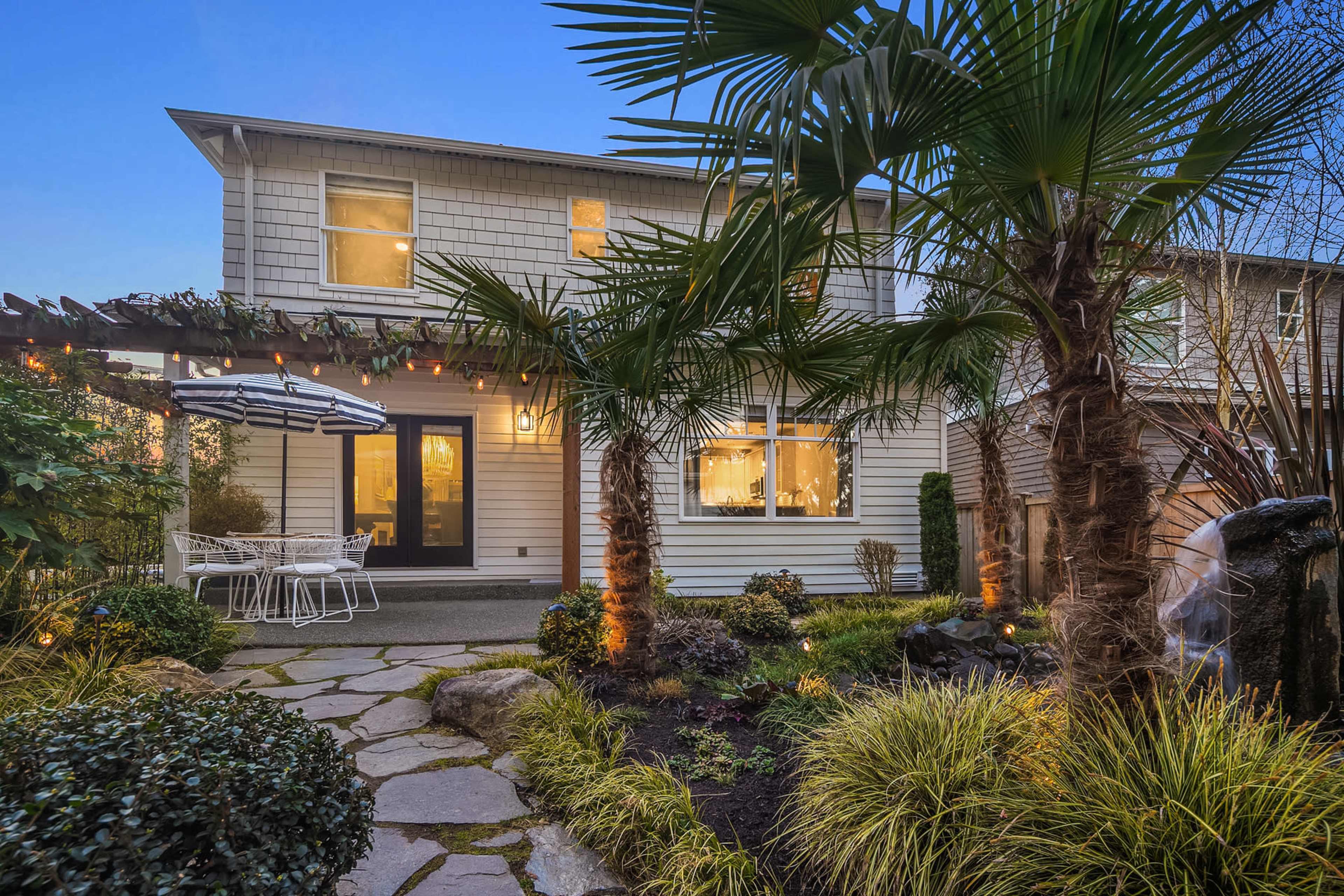A two-story house with a patio and dining area is surrounded by palm trees and landscaped greenery.