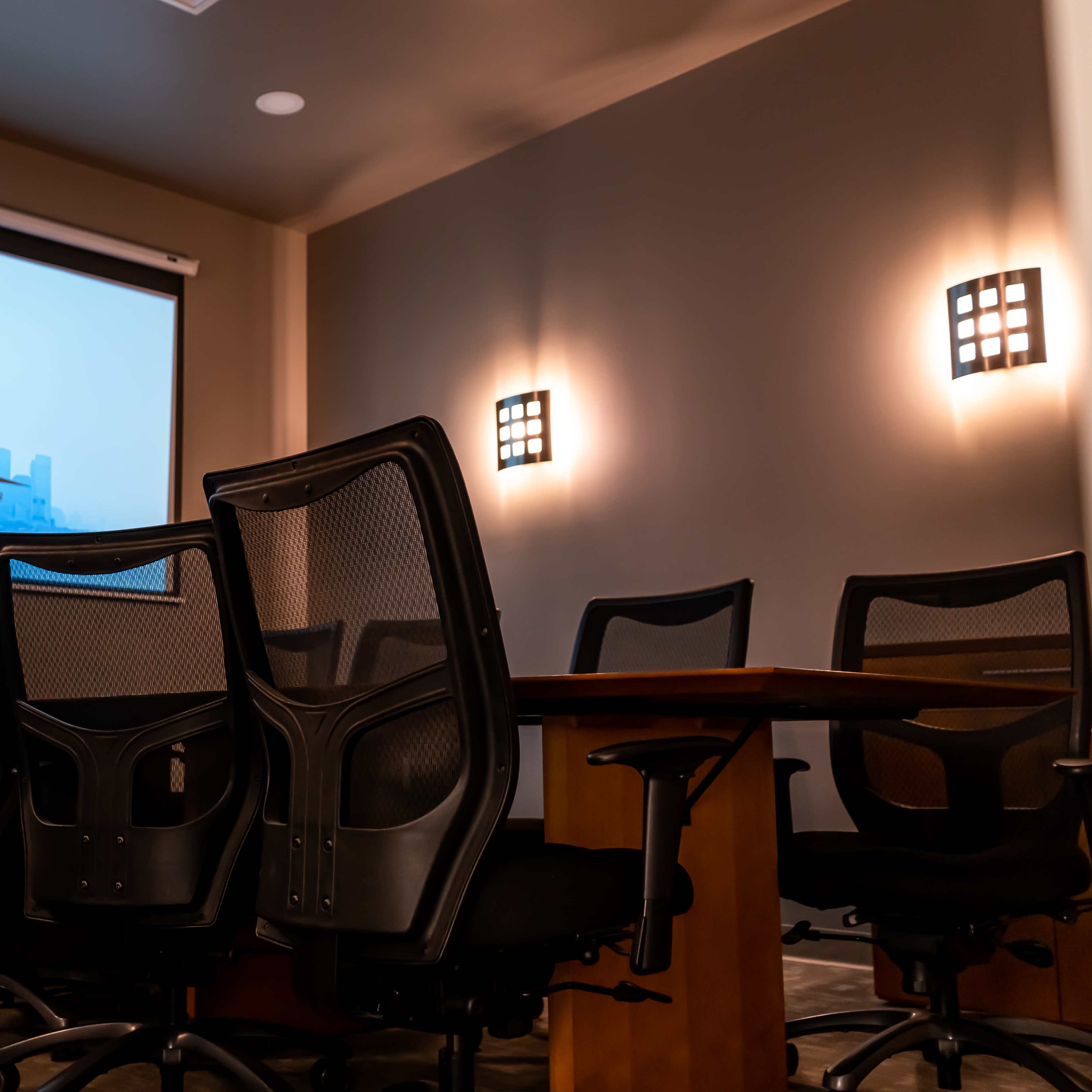 A conference room with a wooden table surrounded by black office chairs, and a screen displaying a city skyline in the background.