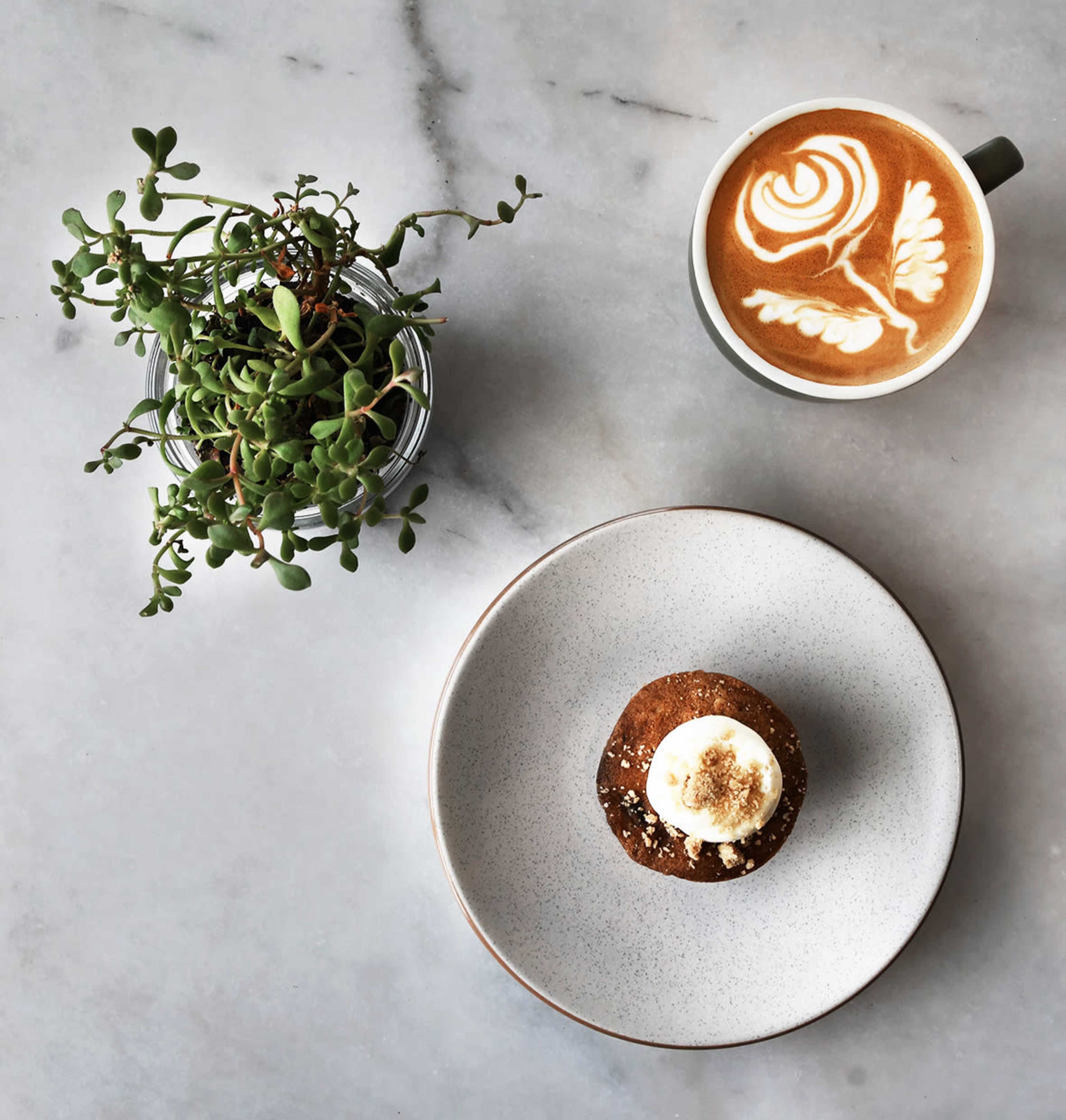 A muffin topped with frosting sits on a plate next to a cup of latte with a floral design, accompanied by a small potted plant on a marble surface.