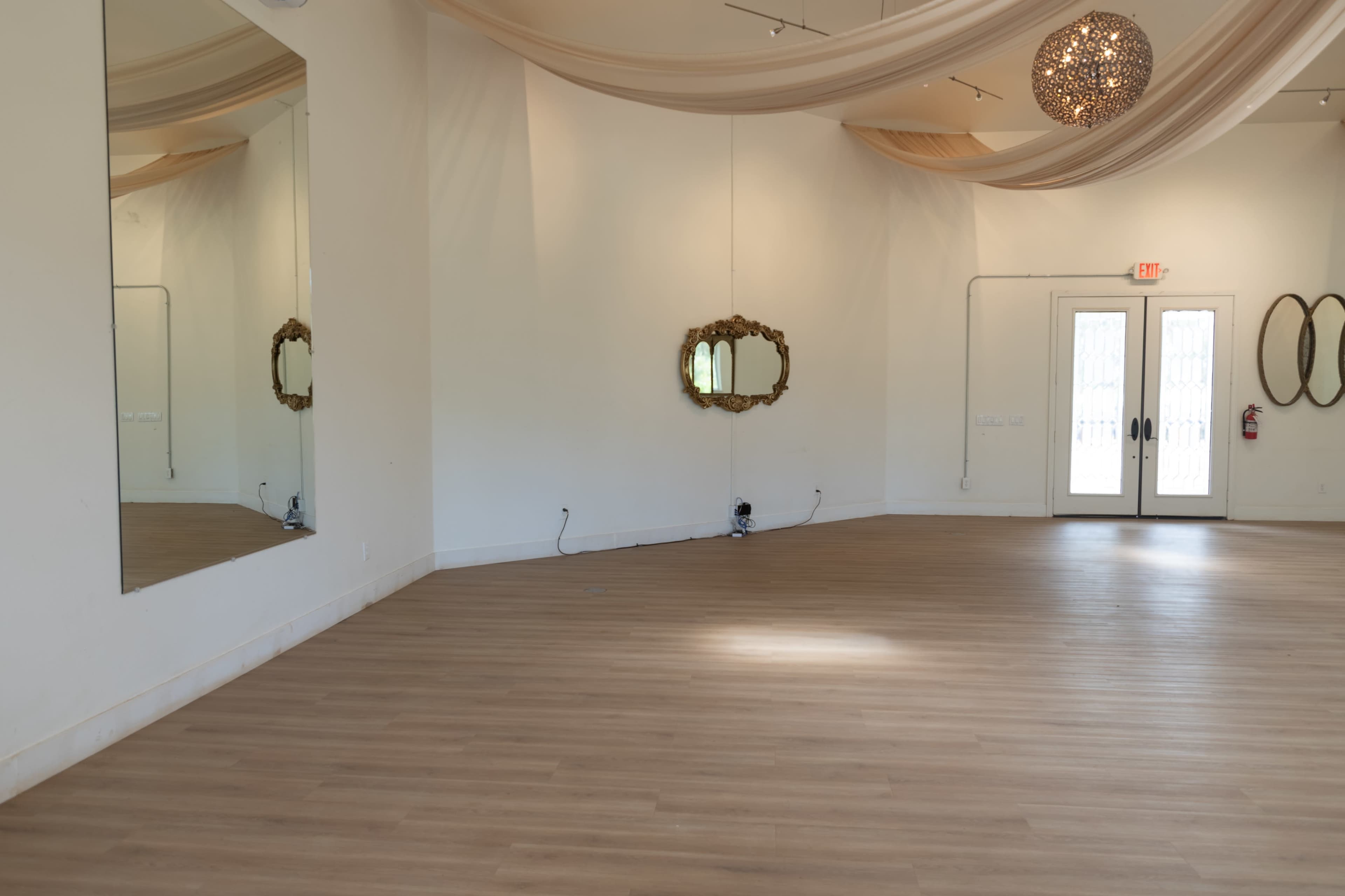 The image shows an empty room with wooden flooring, a large mirror on one wall, and two decorative mirrors hanging on the opposite wall, along with double doors leading outside.
