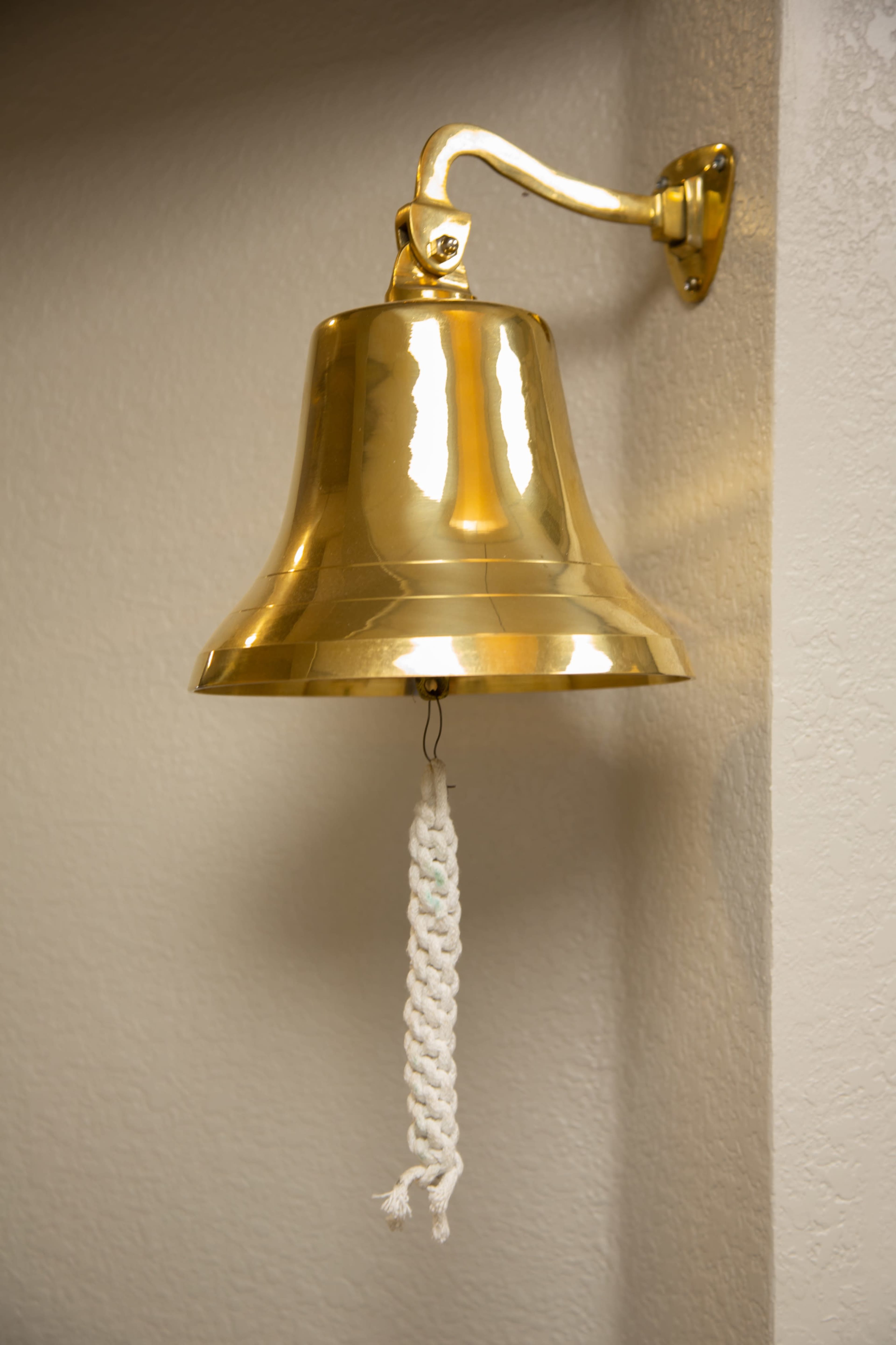 A polished brass bell is mounted on a wall, with a braided white pull cord hanging from its center.
