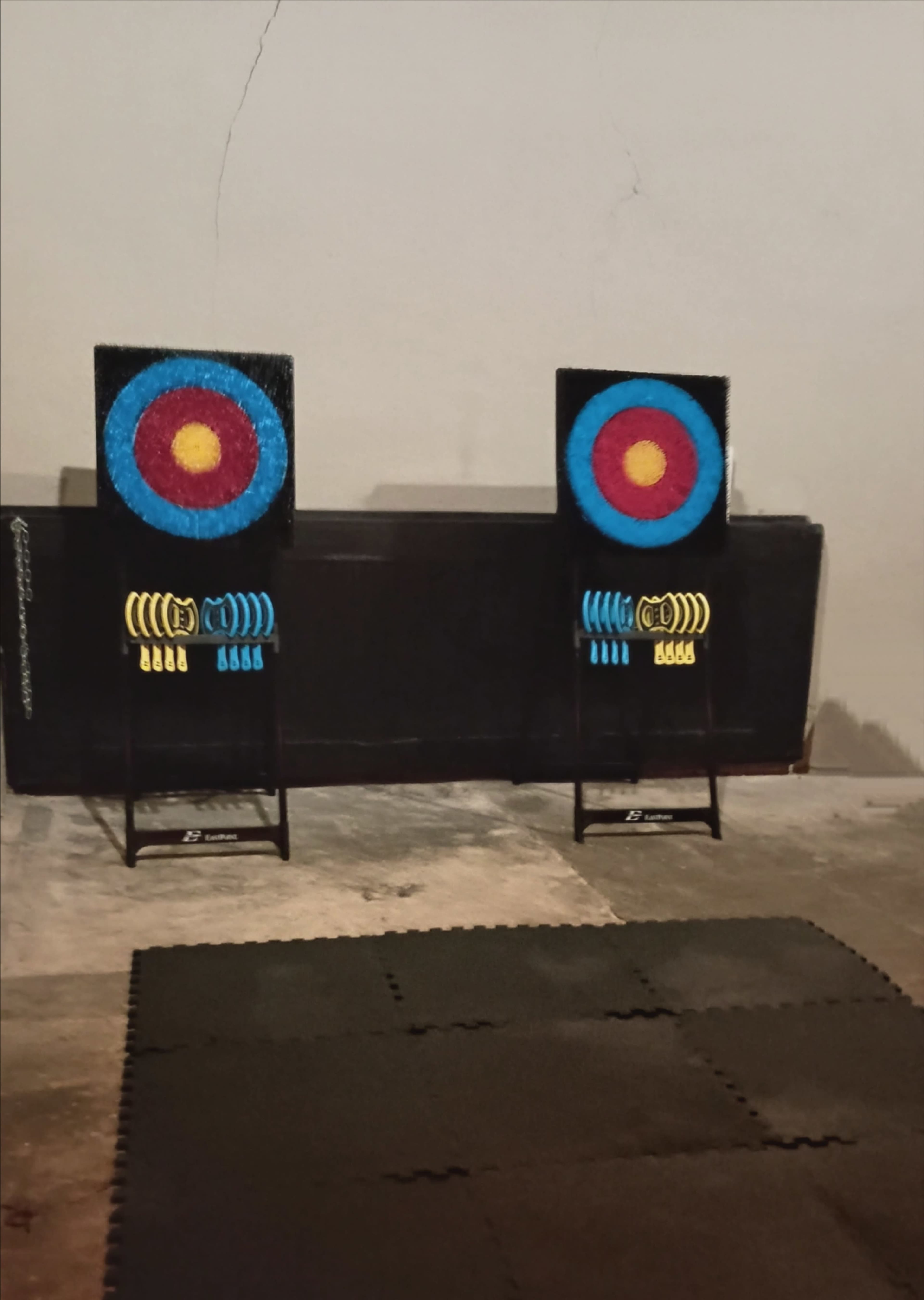 The image shows two colorful archery targets positioned on black stands against a plain wall, with a padded mat on the floor in front of them.