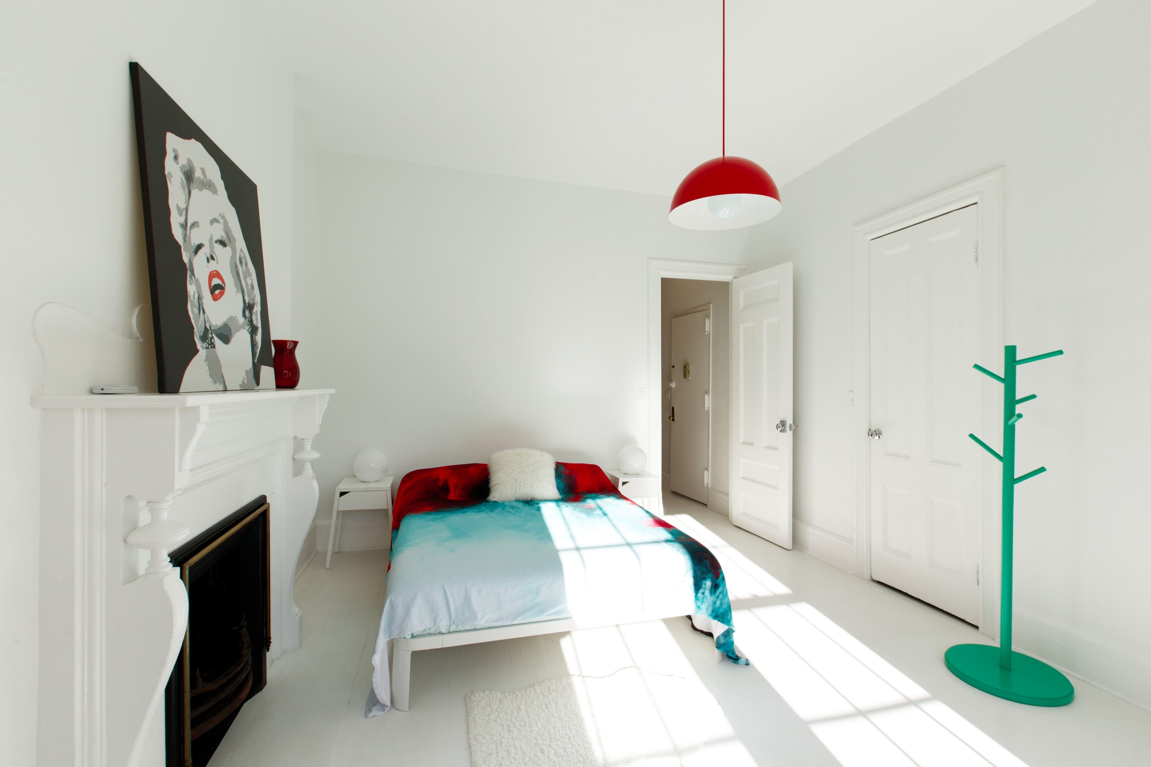A minimalist bedroom featuring a bed with a red and blue blanket, a Marilyn Monroe artwork on the wall, a red pendant light, and a green coat rack.