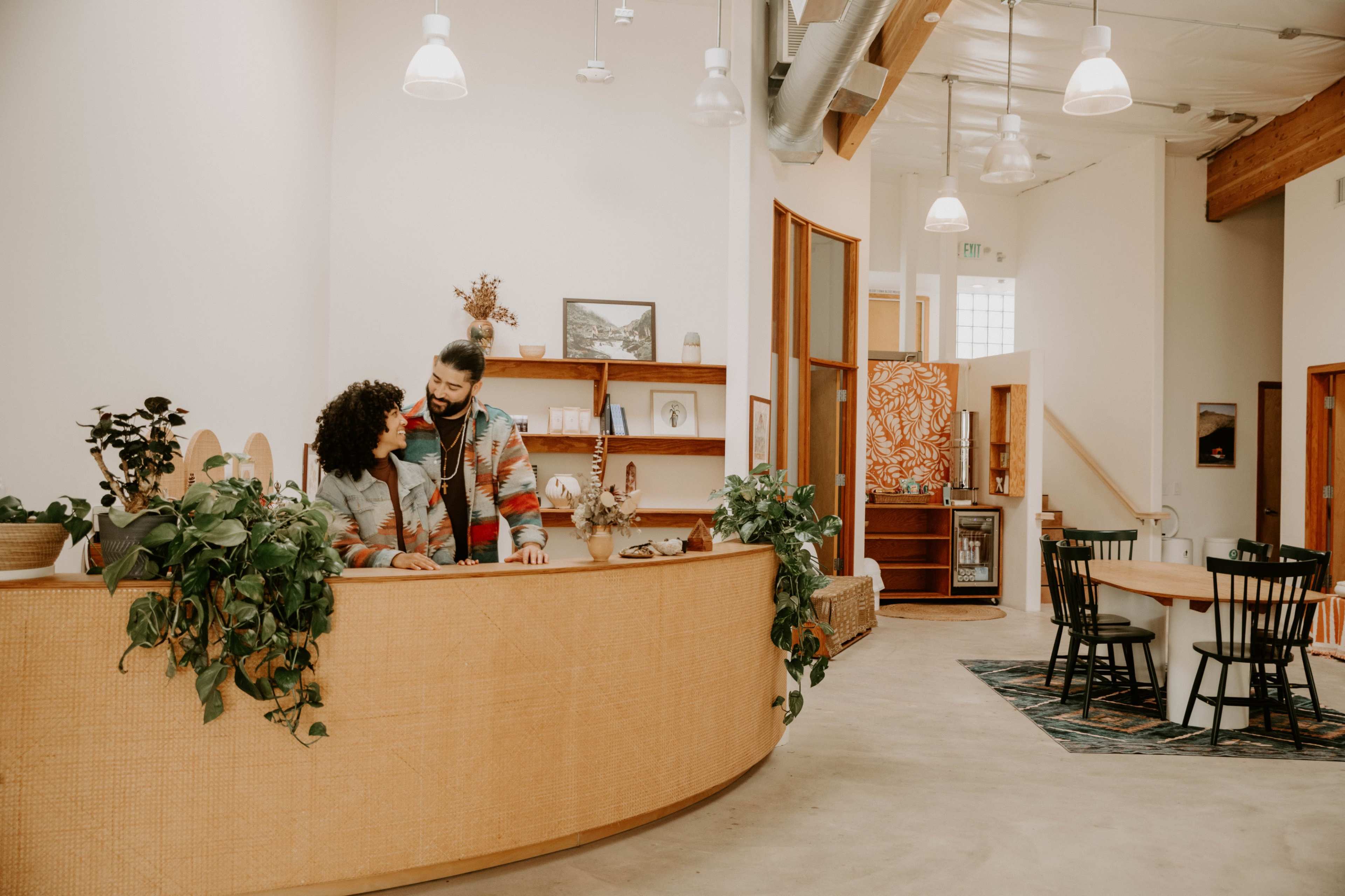 A couple stands together at a curved reception desk surrounded by plants in a spacious, modern interior with wooden accents.