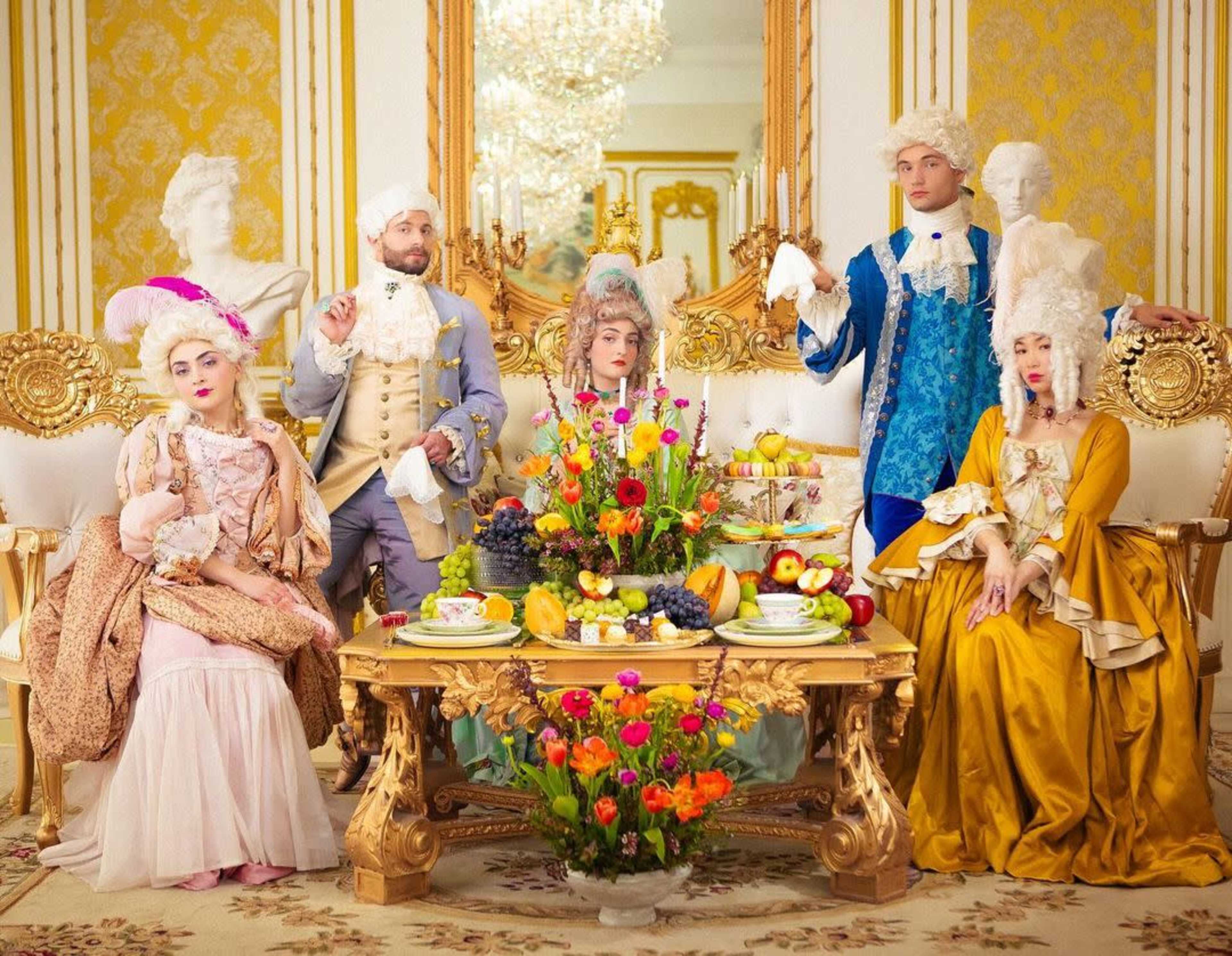A group of elegantly dressed people in 18th-century attire pose around a lavish table filled with fruit and flowers in an ornate interior.