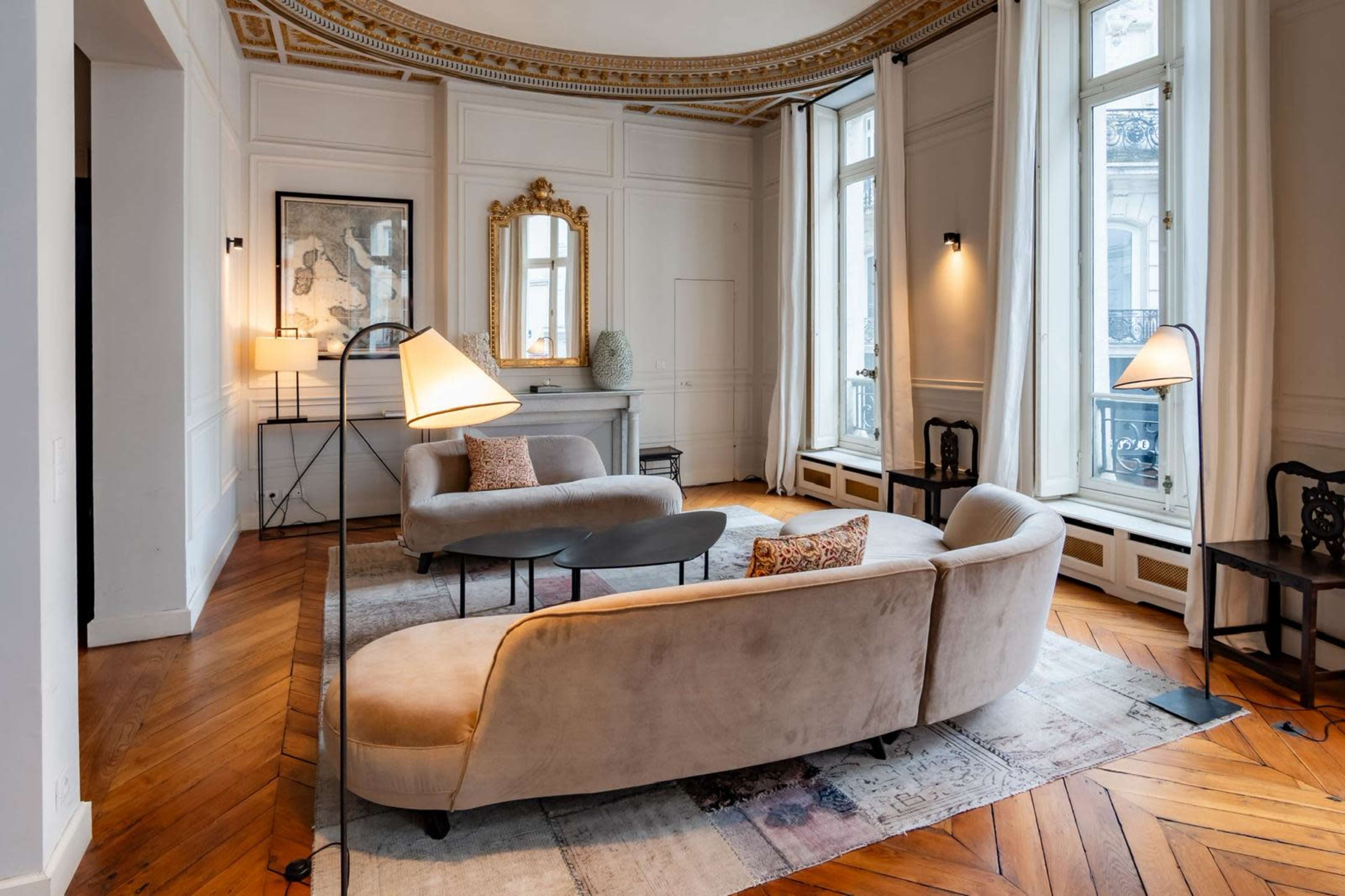 The image shows a stylish living room with a curved sofa, two modern floor lamps, and elegant decor featuring large windows and wooden flooring.