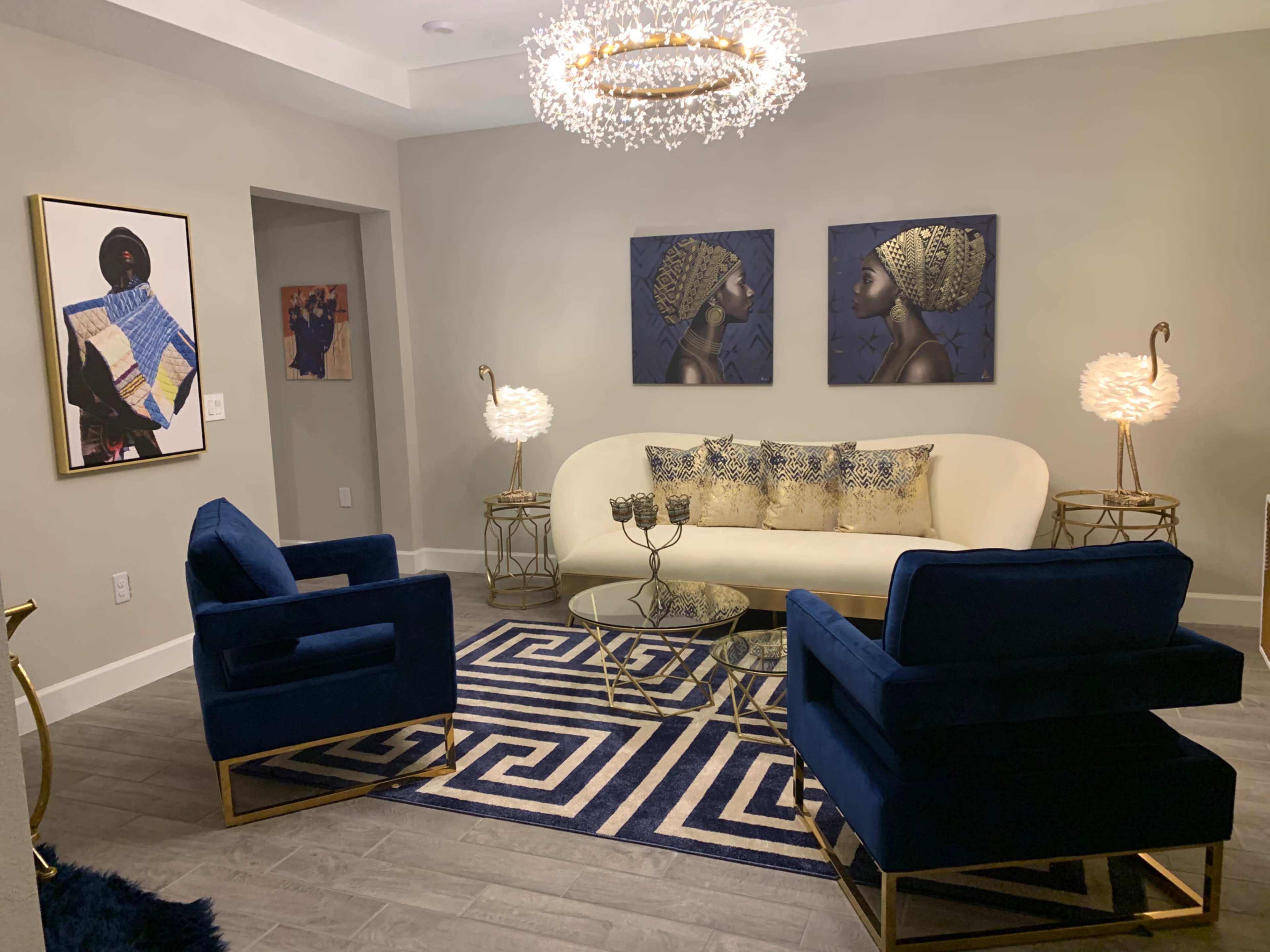 A modern living room featuring a white sofa, two blue armchairs, a glass coffee table, and decorative artwork on the walls.