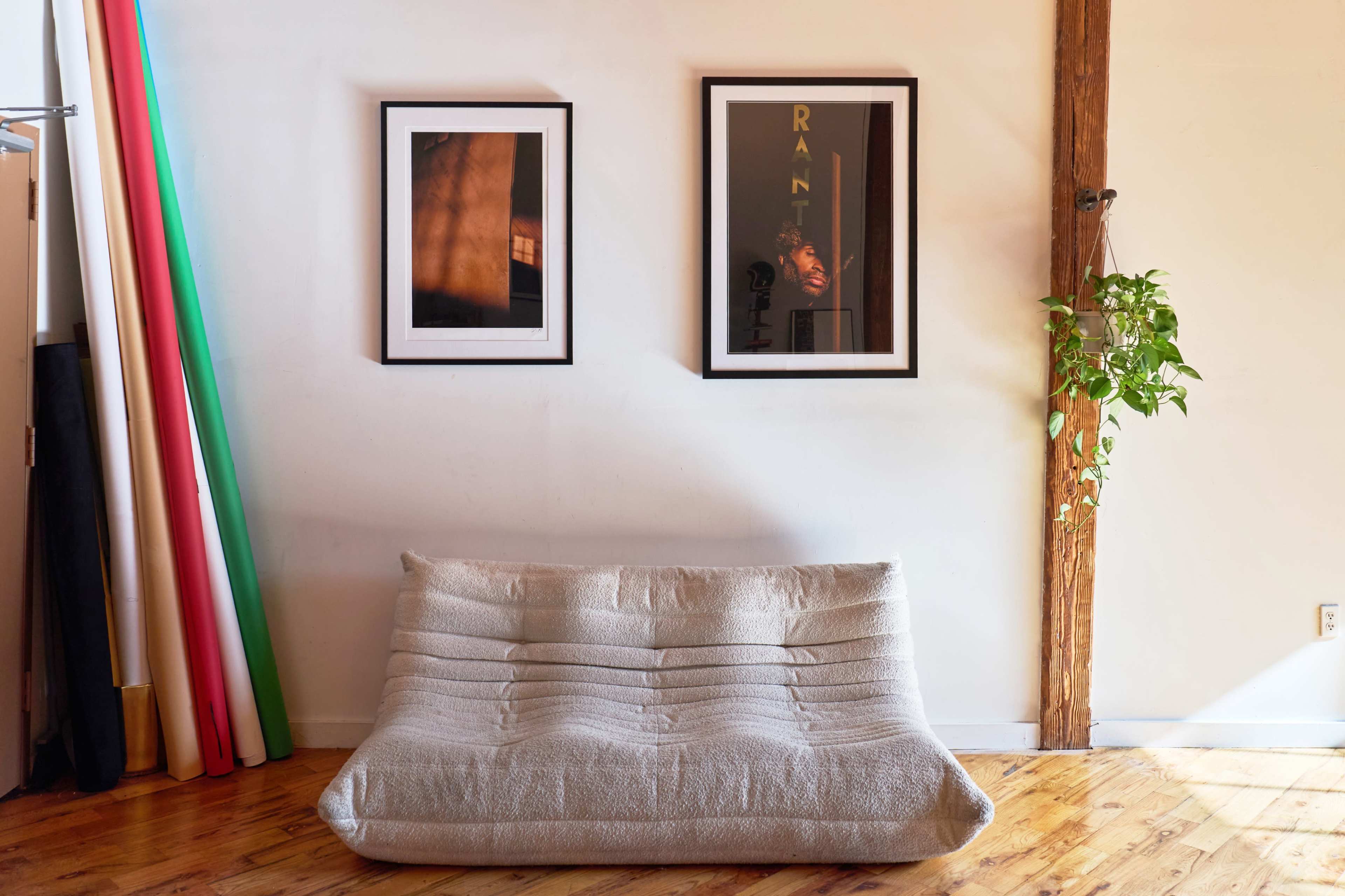 A low, gray fabric sofa is positioned against a wall adorned with two framed pictures, while vibrant colored rolls of material are visible to the left.