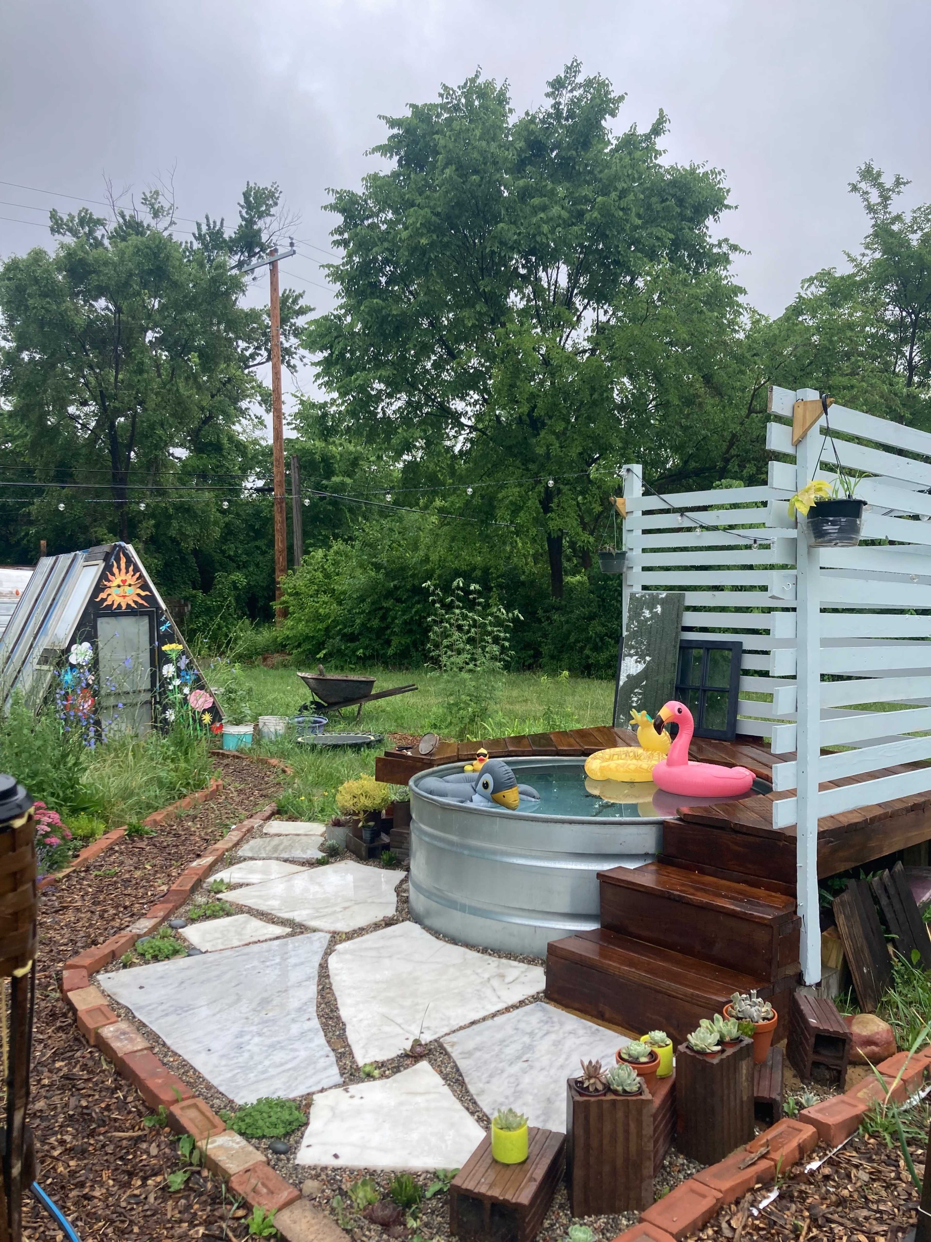 A garden area featuring a galvanized steel water trough, surrounded by stepping stones and wooden decking, with decorative rubber ducks and plants nearby.