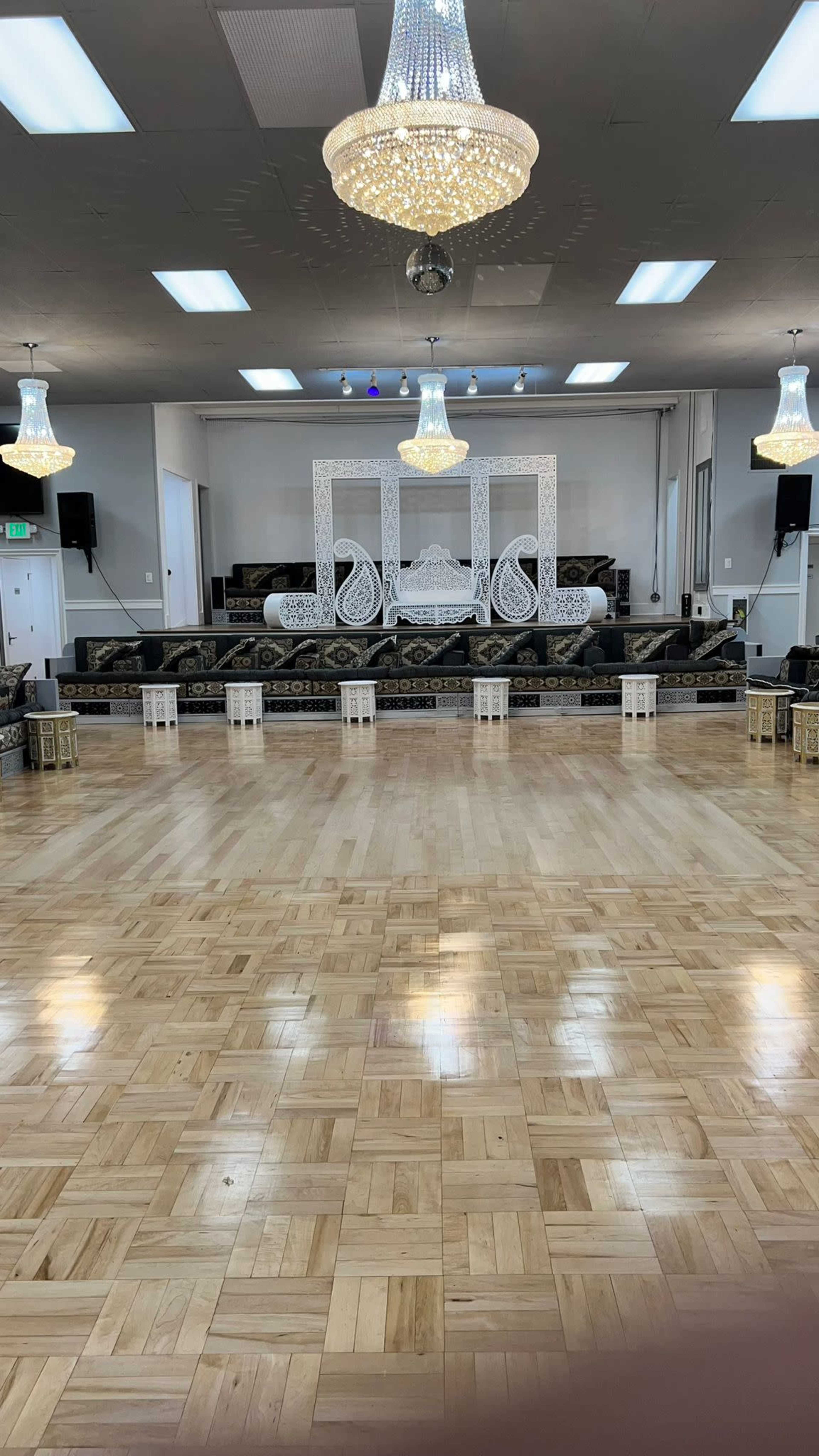 The image shows a spacious event hall with a wooden floor, illuminated by chandeliers, featuring a decorated stage at the front with seating and decorations along the sides.