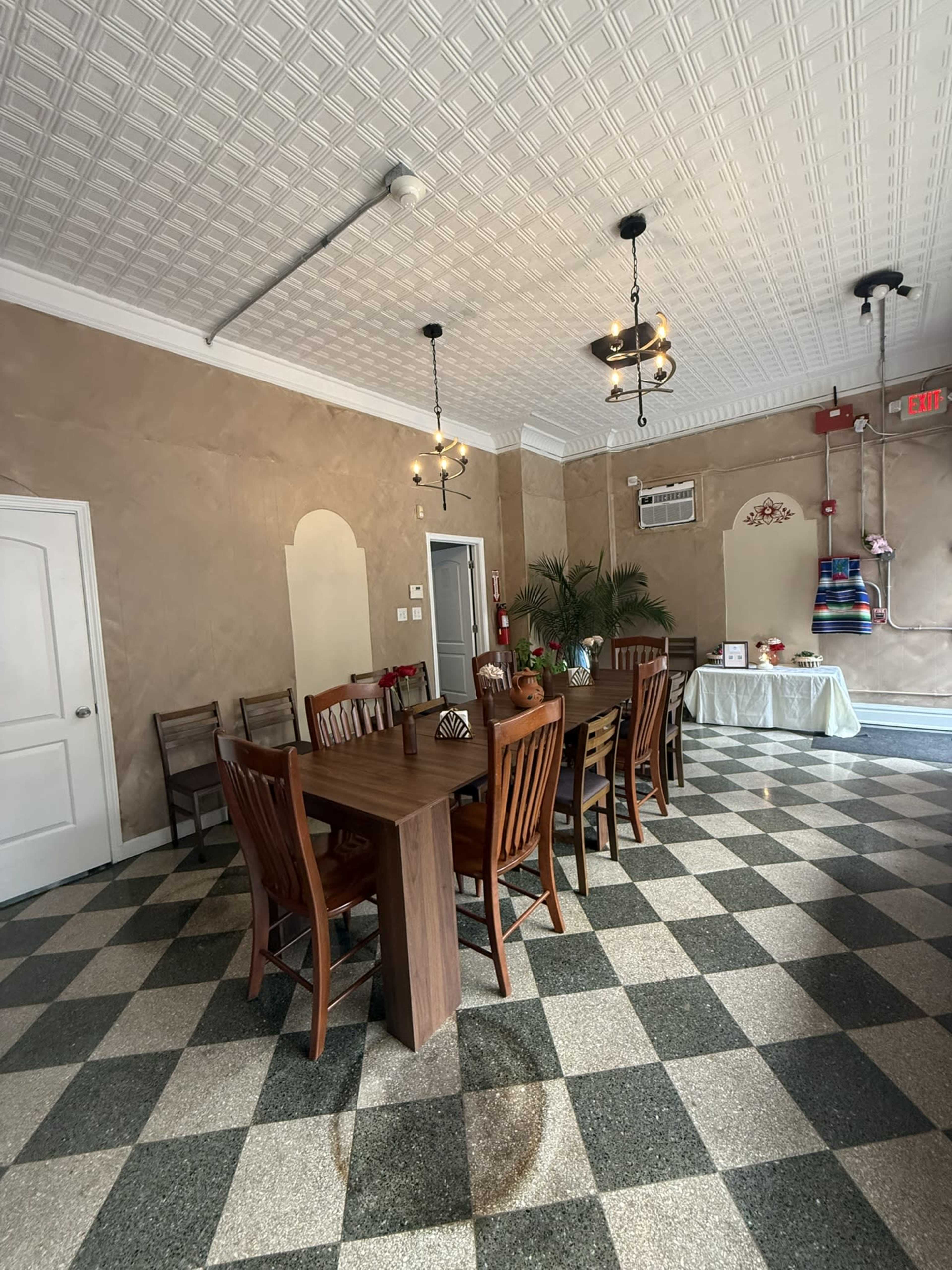 The image shows a spacious dining area featuring a long wooden table surrounded by chairs, with patterned flooring and simple decorative elements on the walls.