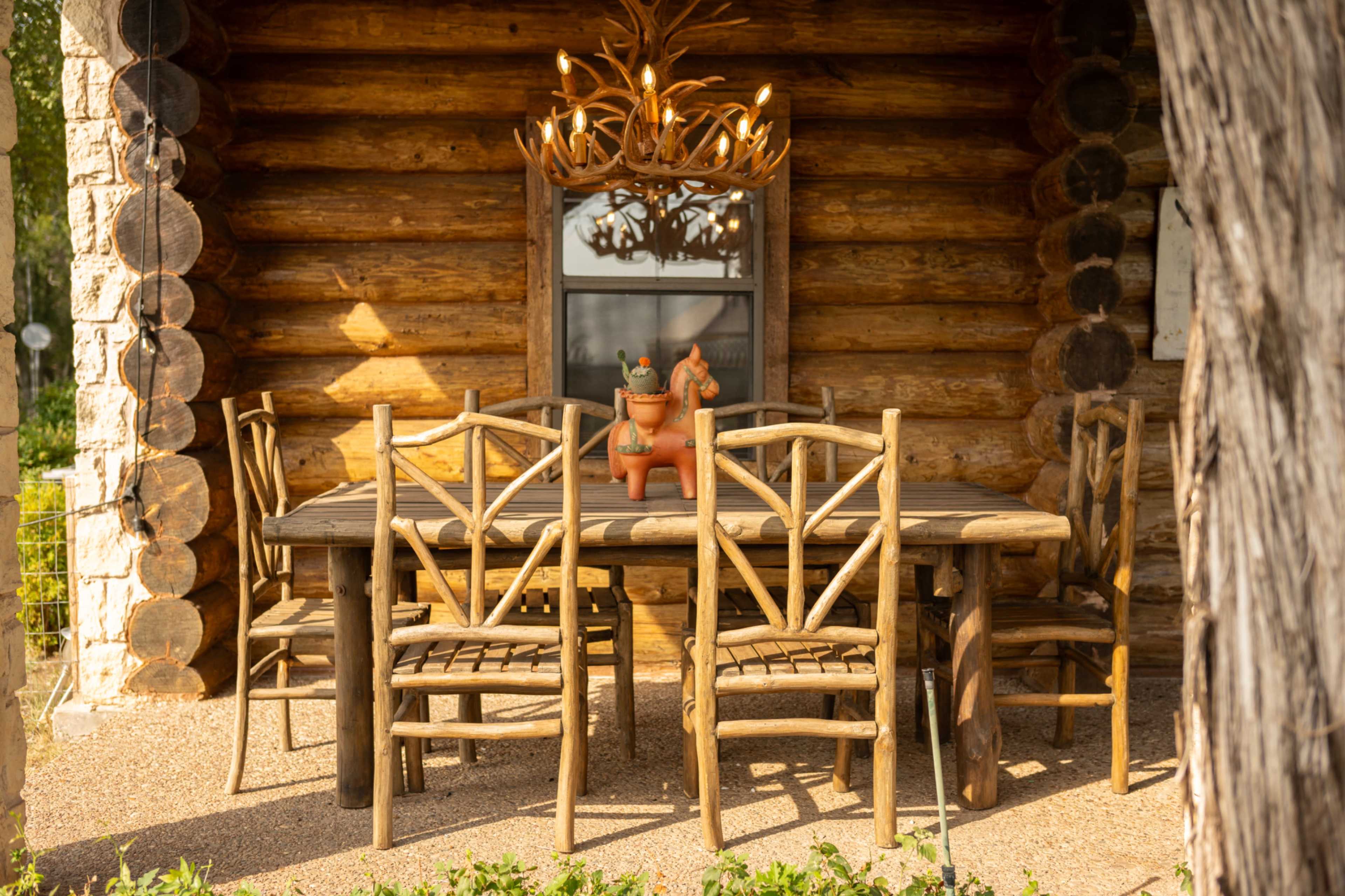 A rustic wooden dining table with matching chairs is set in front of a log cabin, featuring an ornately designed chandelier above.