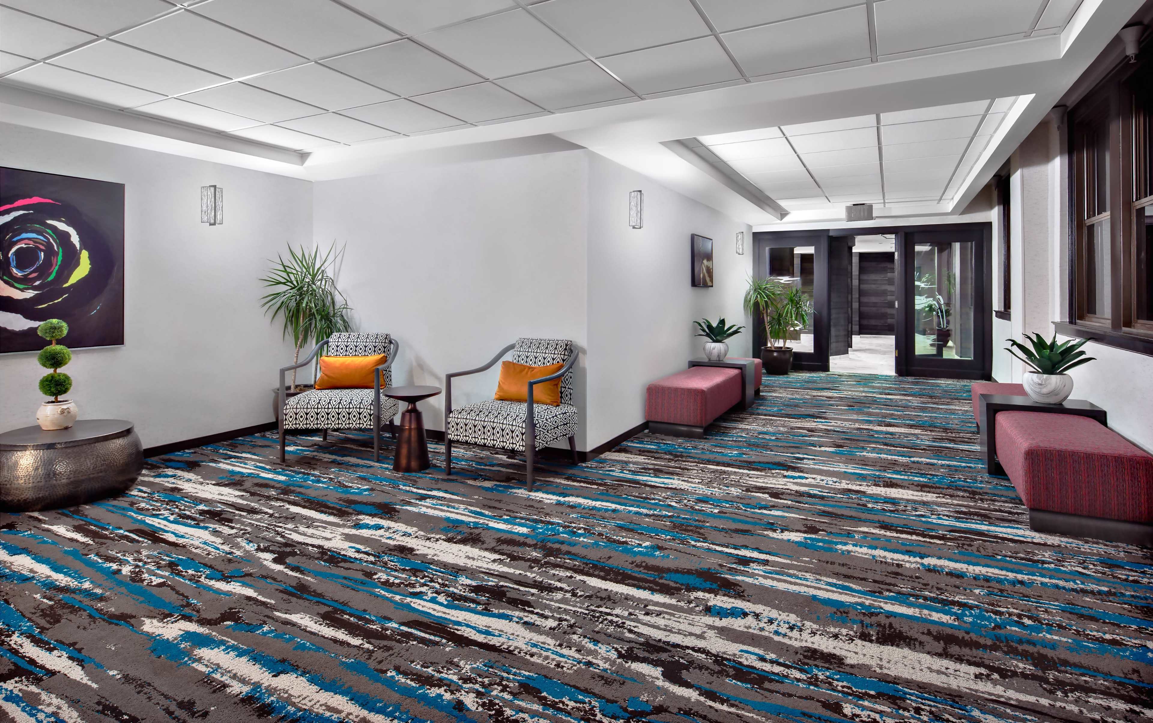 The image shows a modern hallway with patterned carpet, two armchairs with orange cushions, a small table, and large windows at one end.
