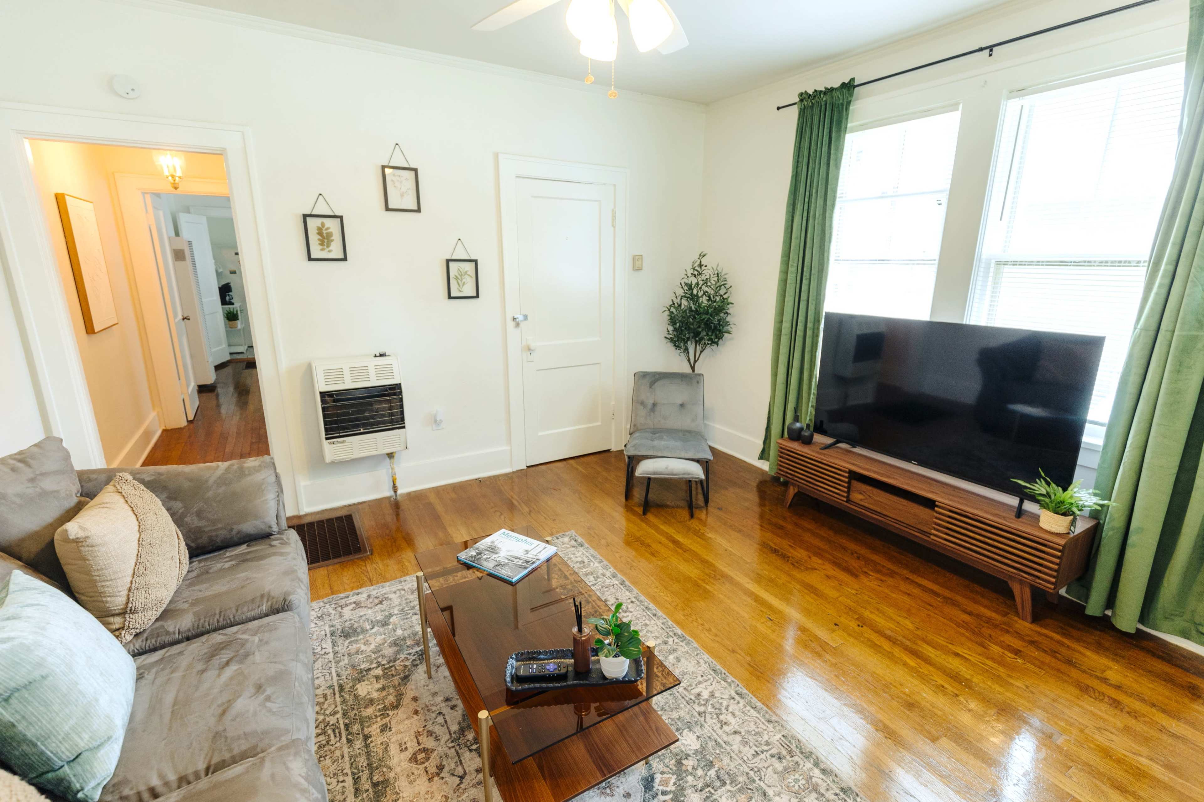 A living room with a sofa, a coffee table, a television stand, and large windows dressed with green curtains.