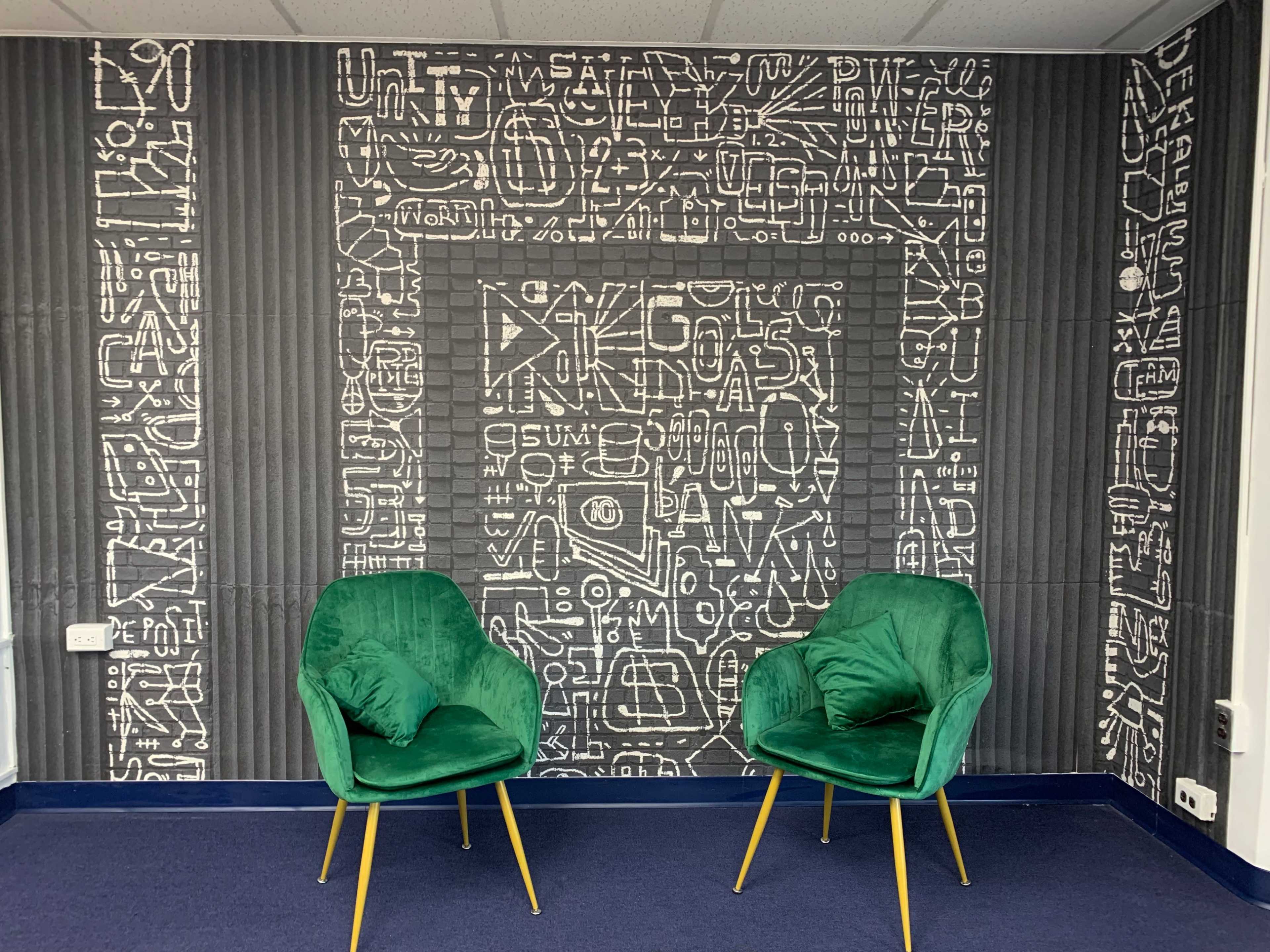 A room with a patterned wall covered in white text and symbols, featuring two green velvet chairs with gold legs positioned in front of it.