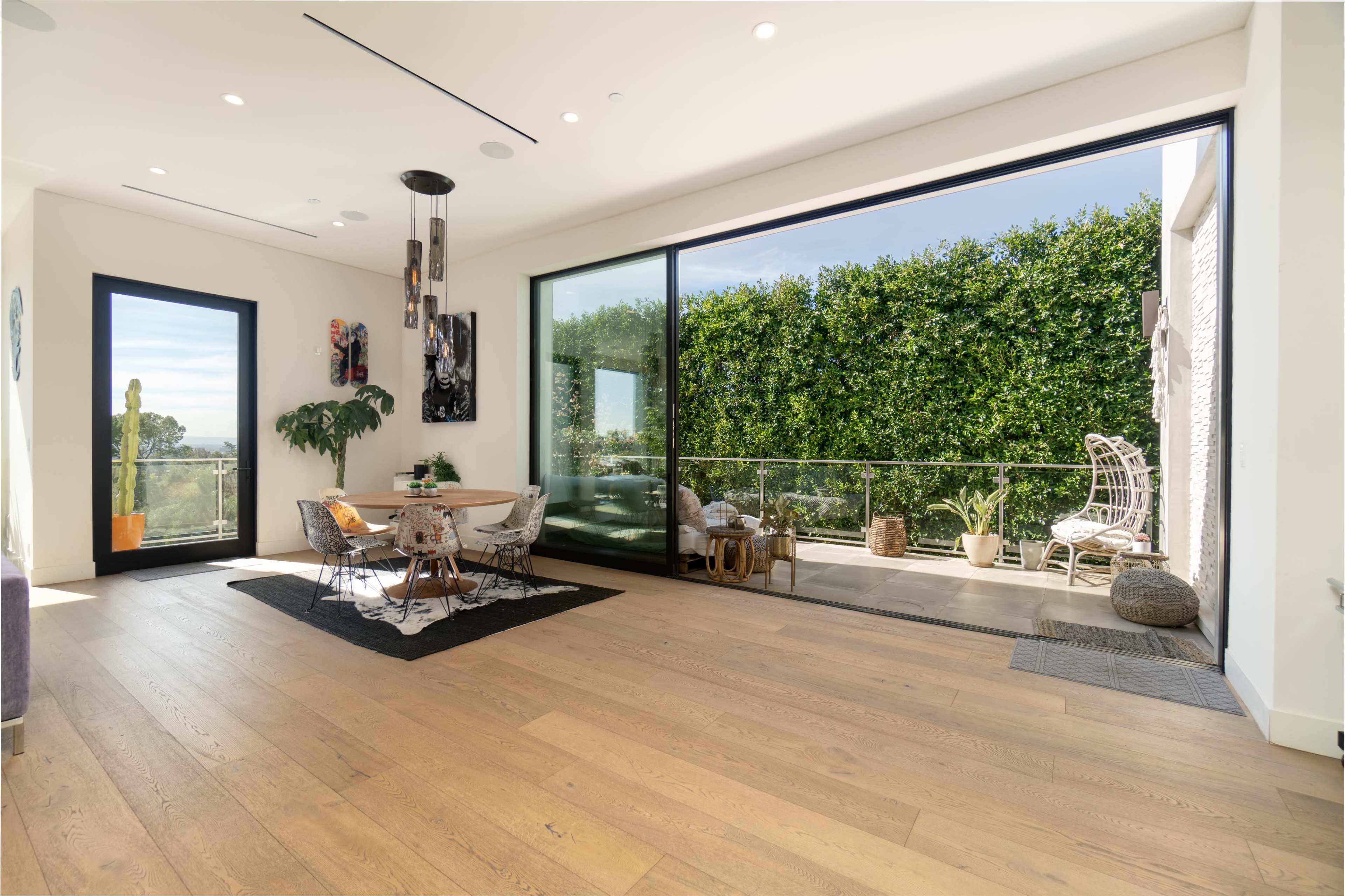 The image shows a spacious living area with large windows overlooking a balcony adorned with plants and outdoor seating.