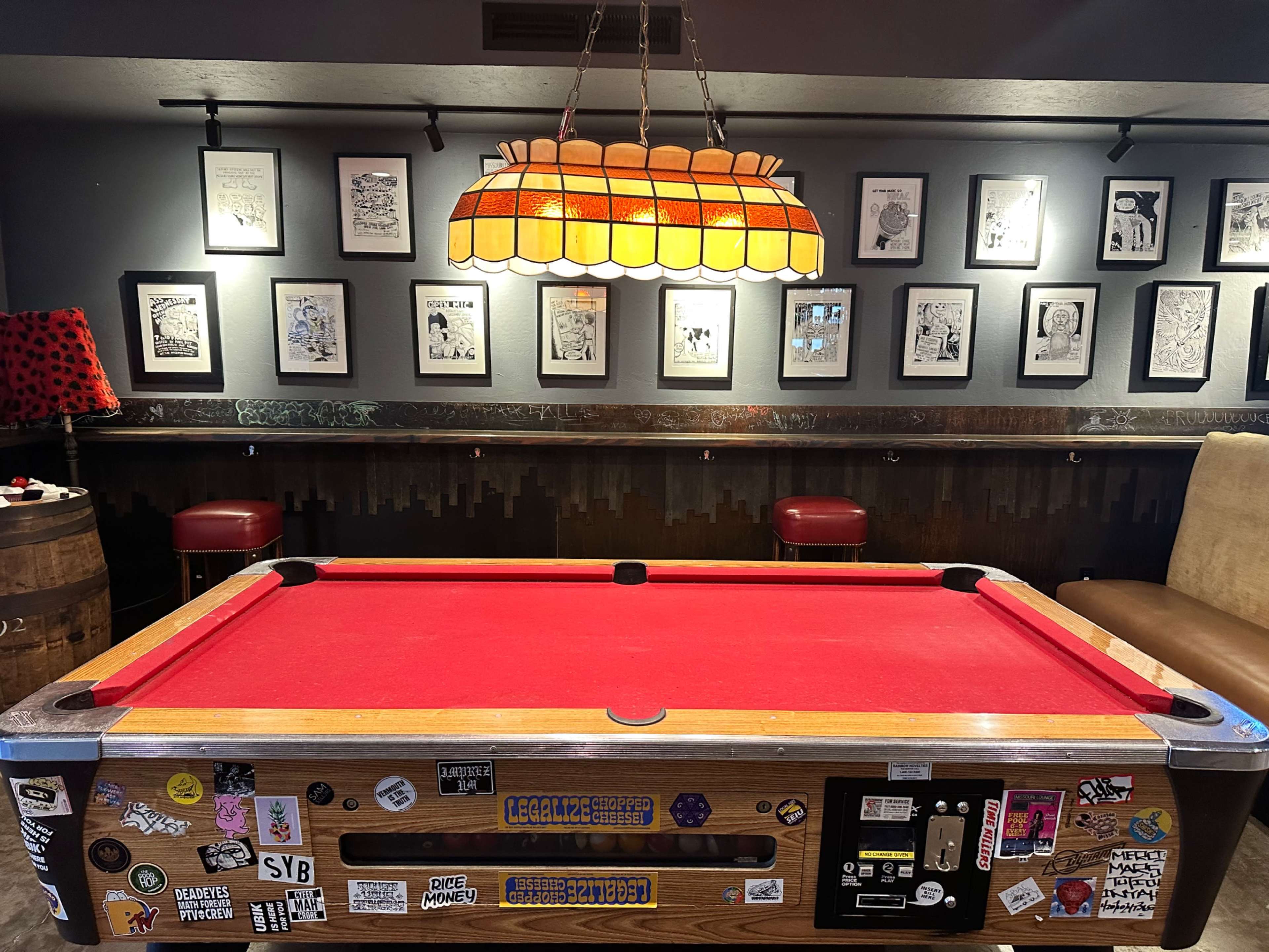 A red pool table with various stickers on its sides is positioned in a dimly lit room adorned with framed artworks on the walls.