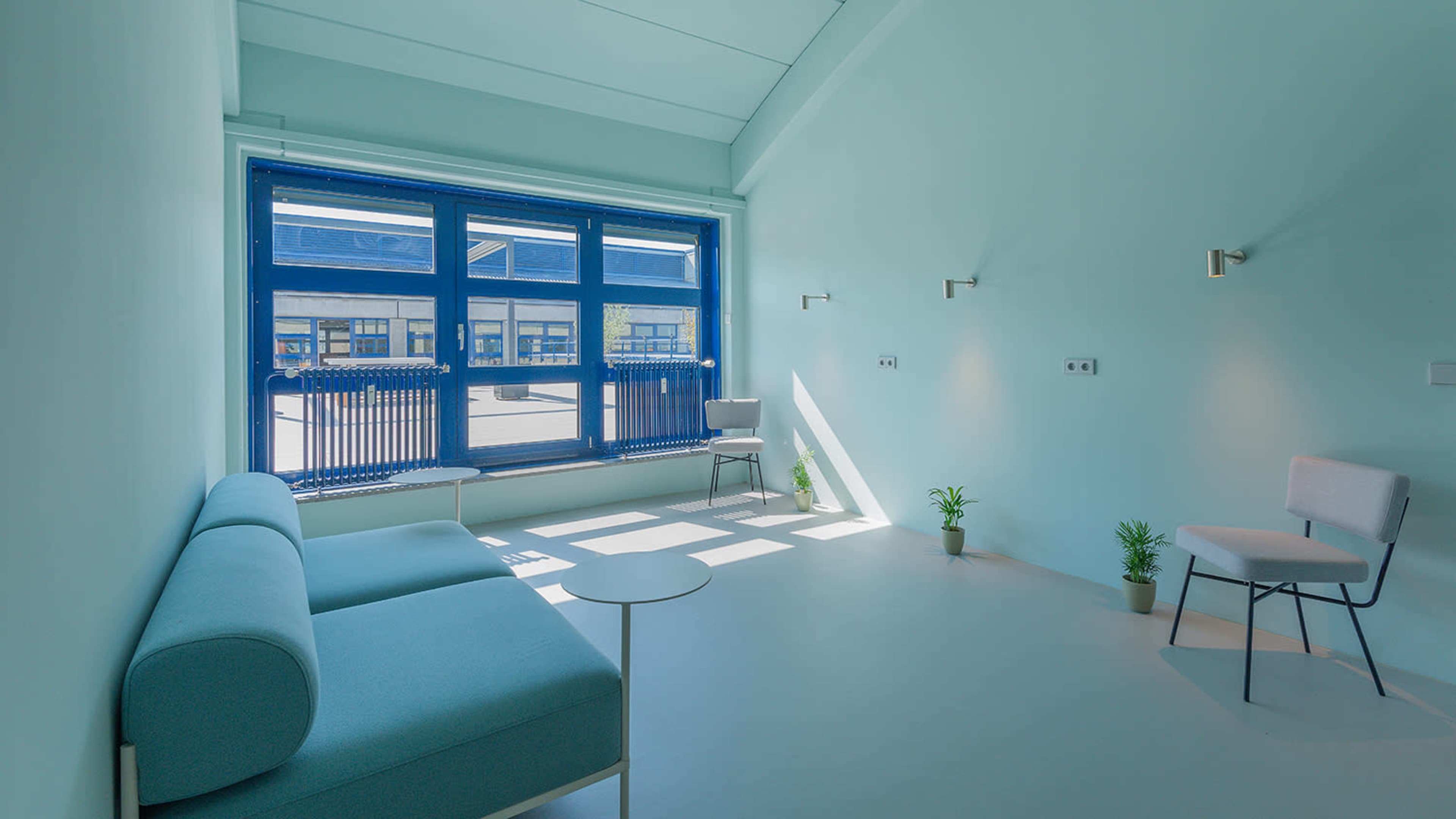 The image shows a minimalist room with light blue walls, a large window with blue frames, a small table, a sofa, and a chair.