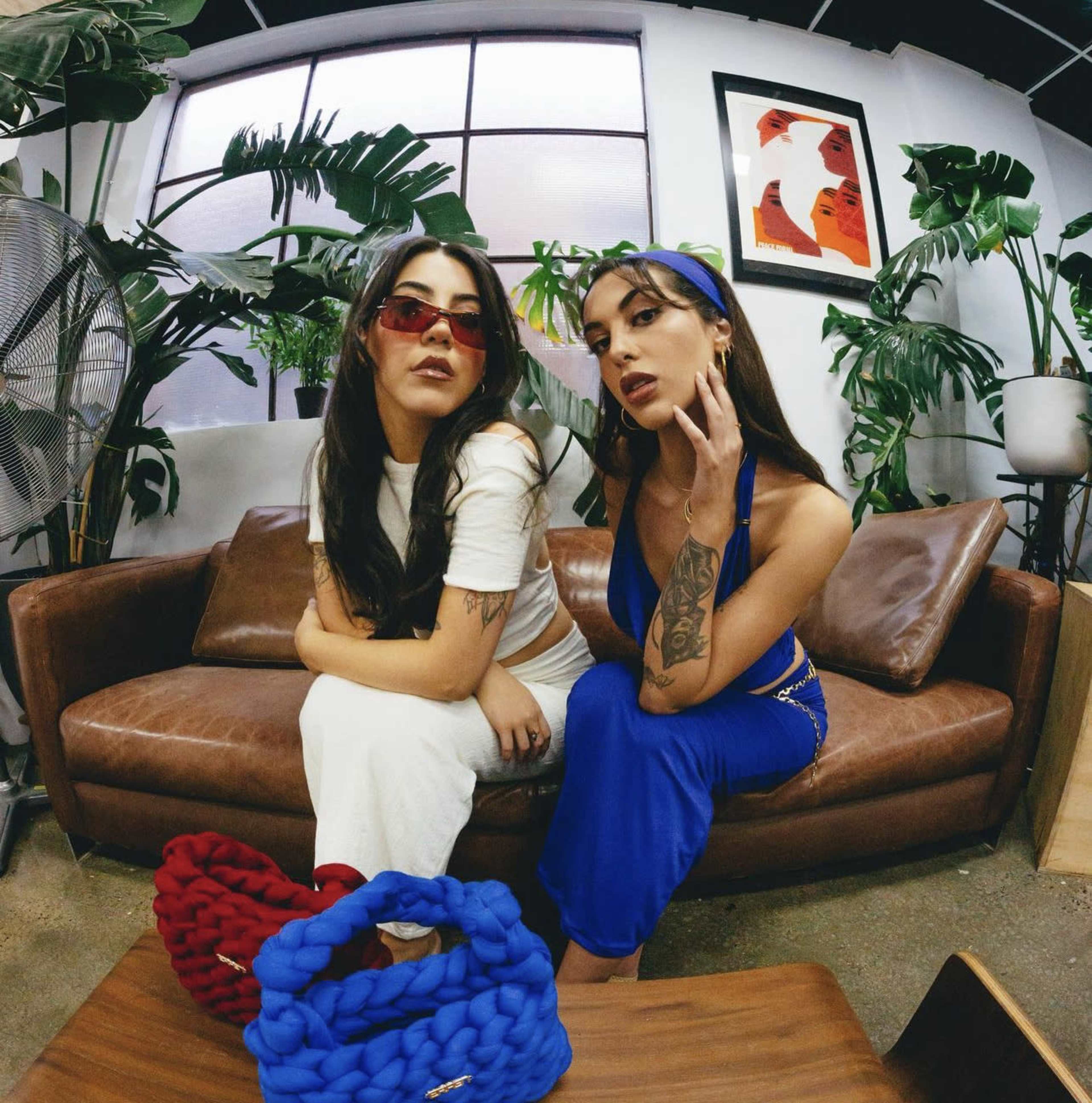 Two women pose on a brown leather sofa in a green, plant-filled room, wearing colorful outfits and accessories.