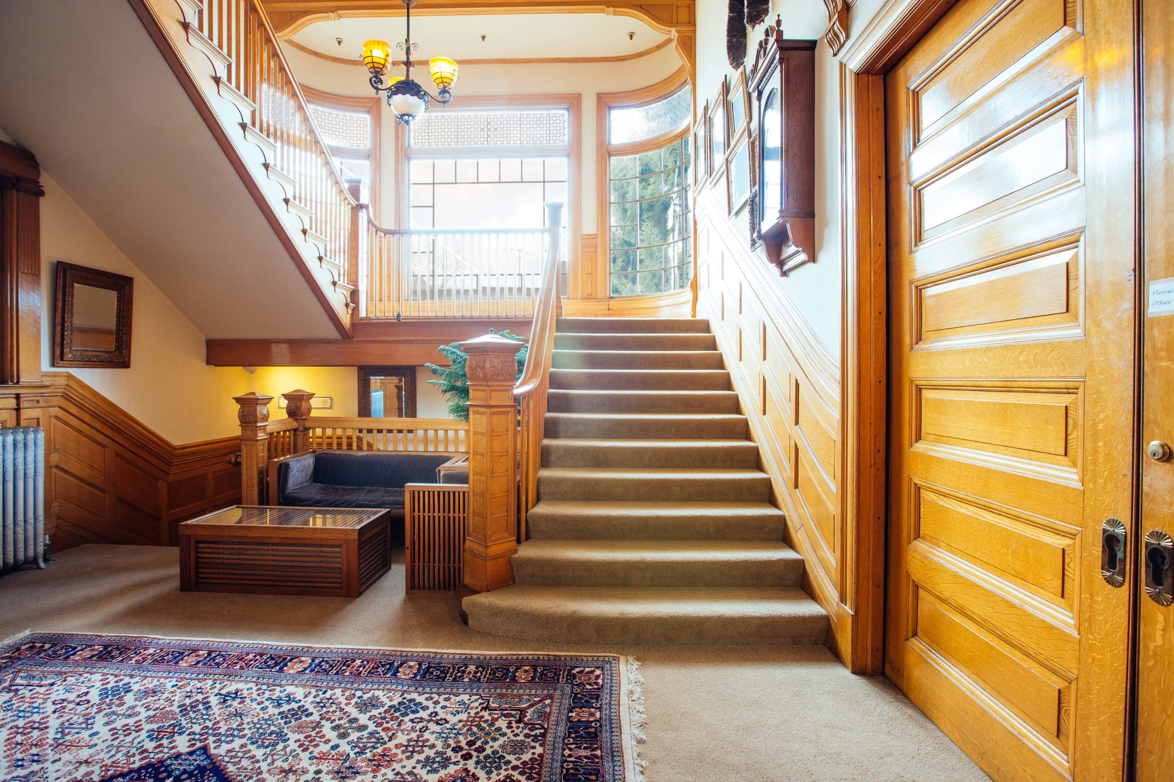 The image shows a stairway in a vintage interior with wooden paneling, a decorative rug on the floor, and light streaming in through windows.