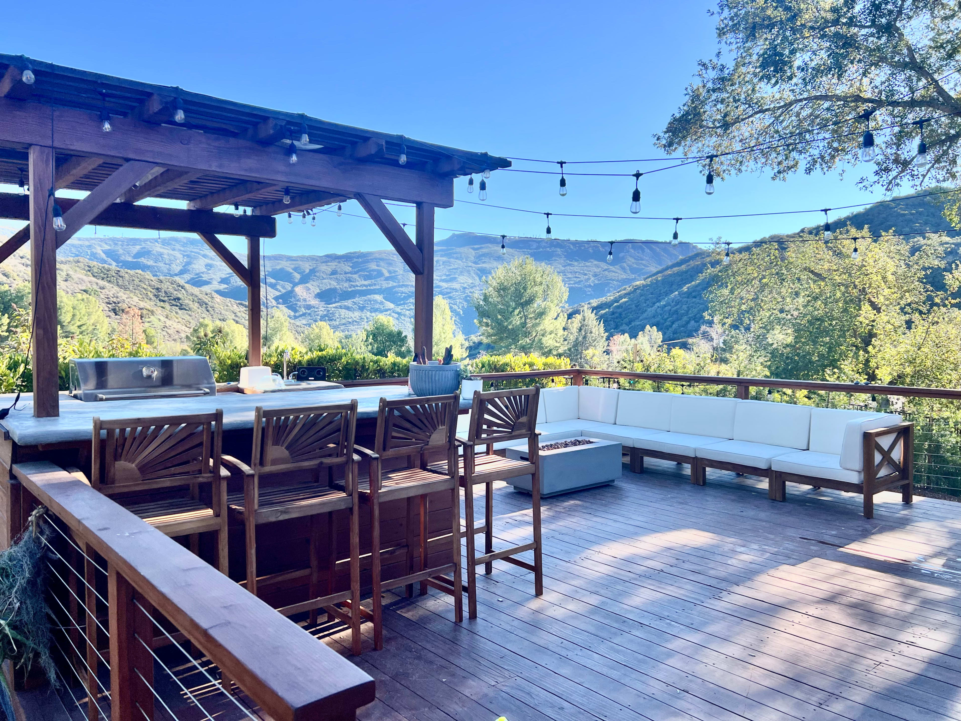 A wooden deck with bar stools, a fire pit, and a seating area, overlooking a panoramic view of mountains and trees under a clear blue sky.