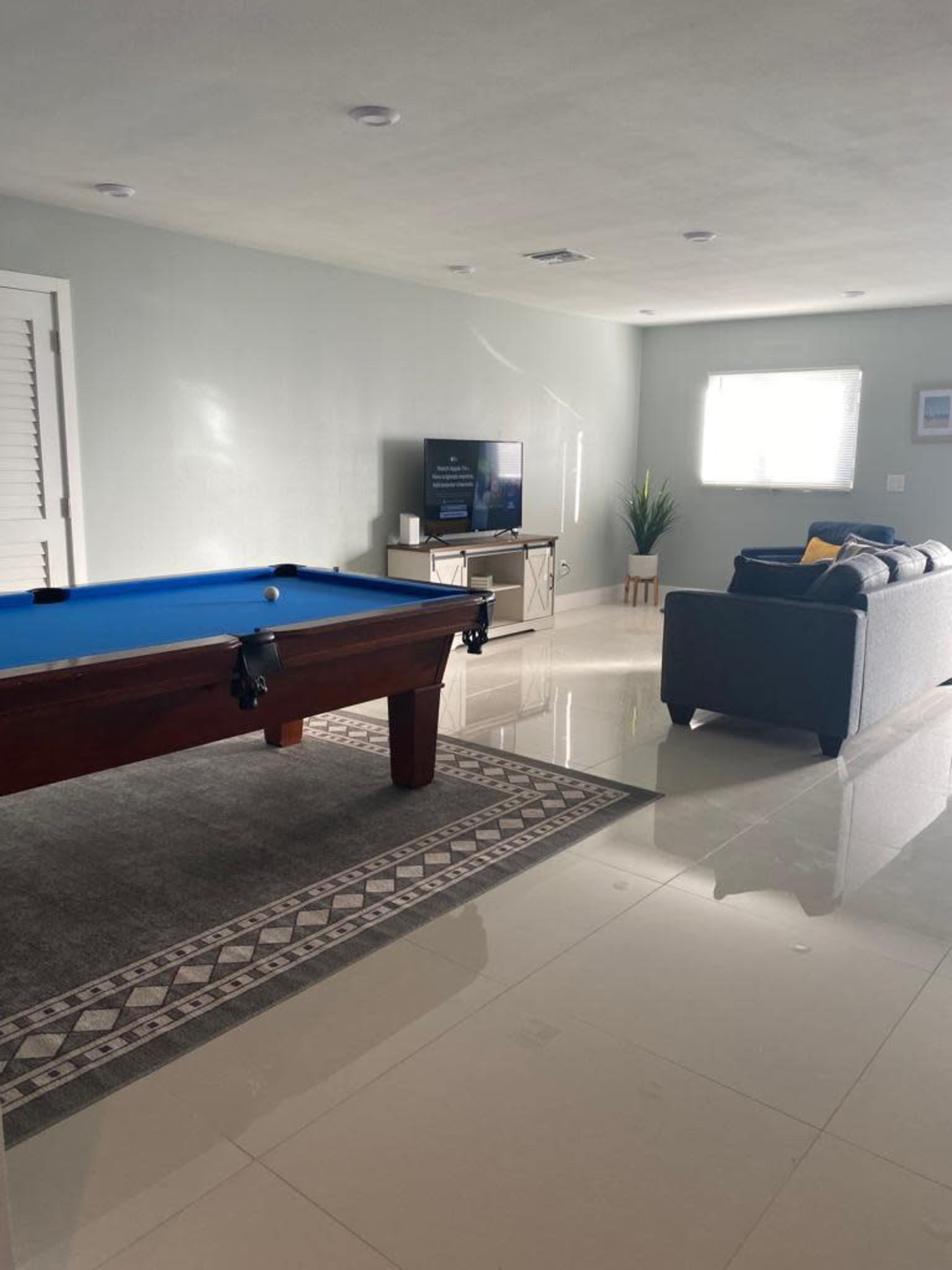 The image shows a room with a blue pool table, a gray sofa, a television on a stand, and a bright window.