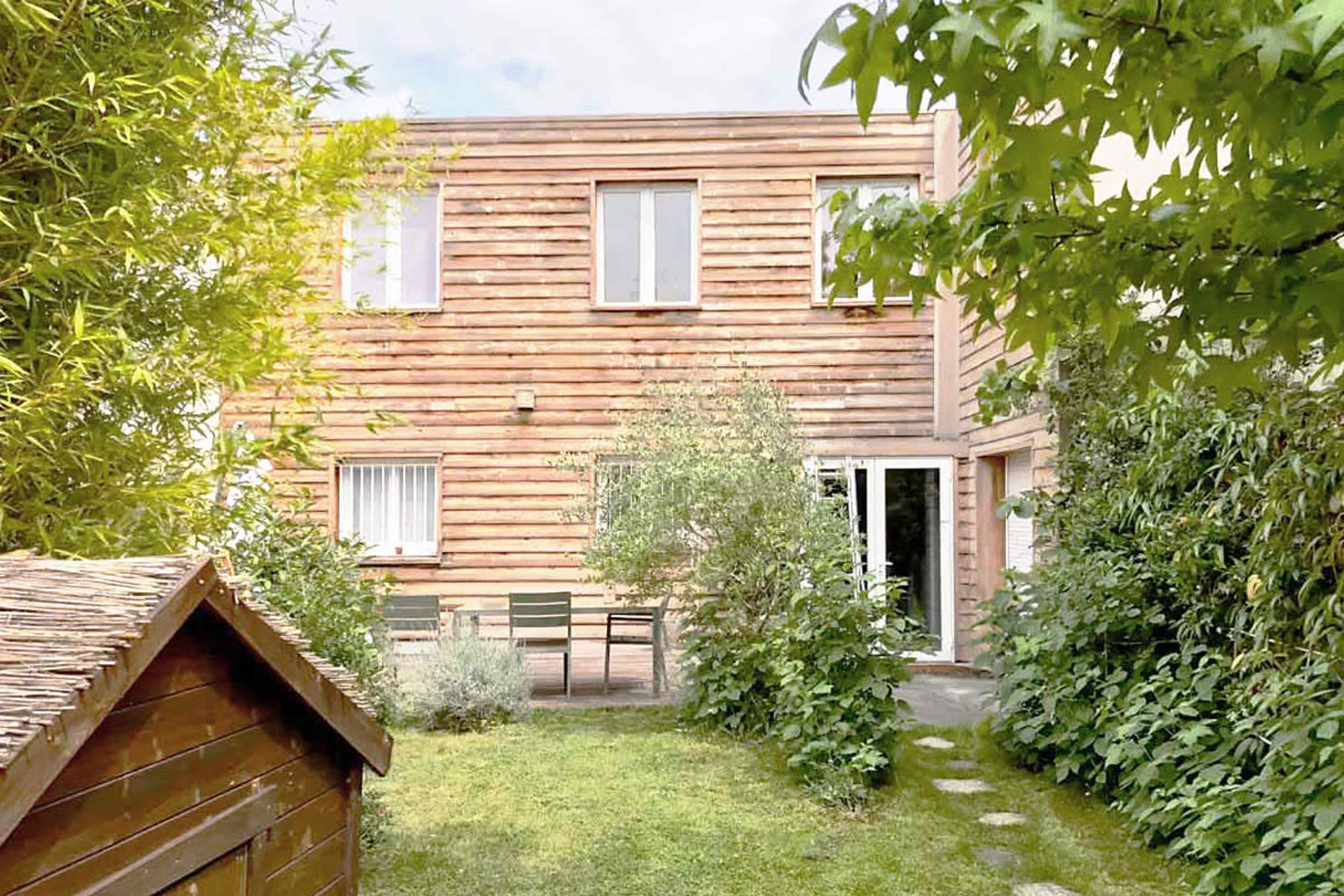 A wooden house with multiple windows is surrounded by greenery and a small garden, featuring a pathway leading to a seating area.