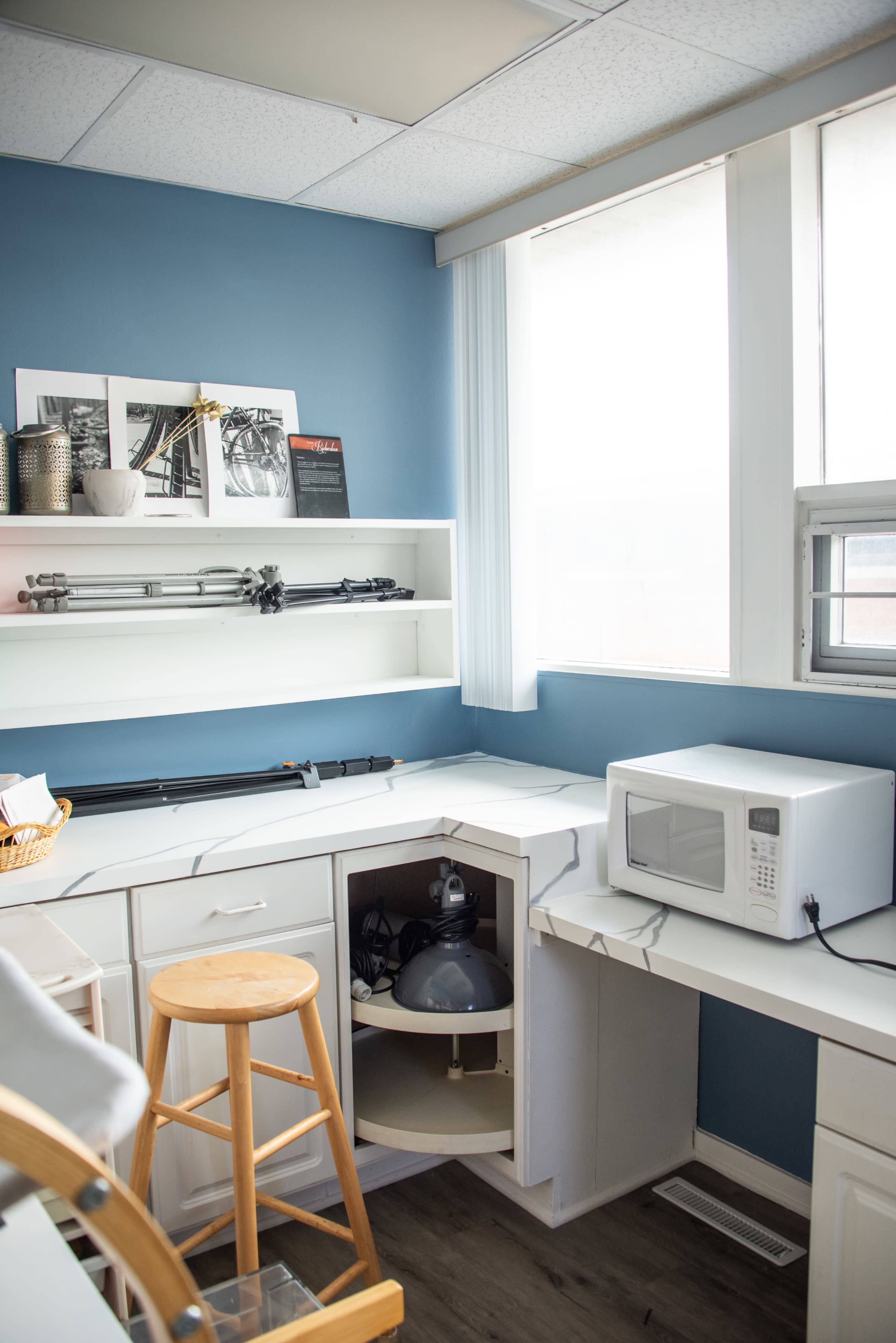 The image shows a tidy corner of a room featuring a microwave, a stool, and shelves displaying framed photographs and kitchen items against a blue wall.