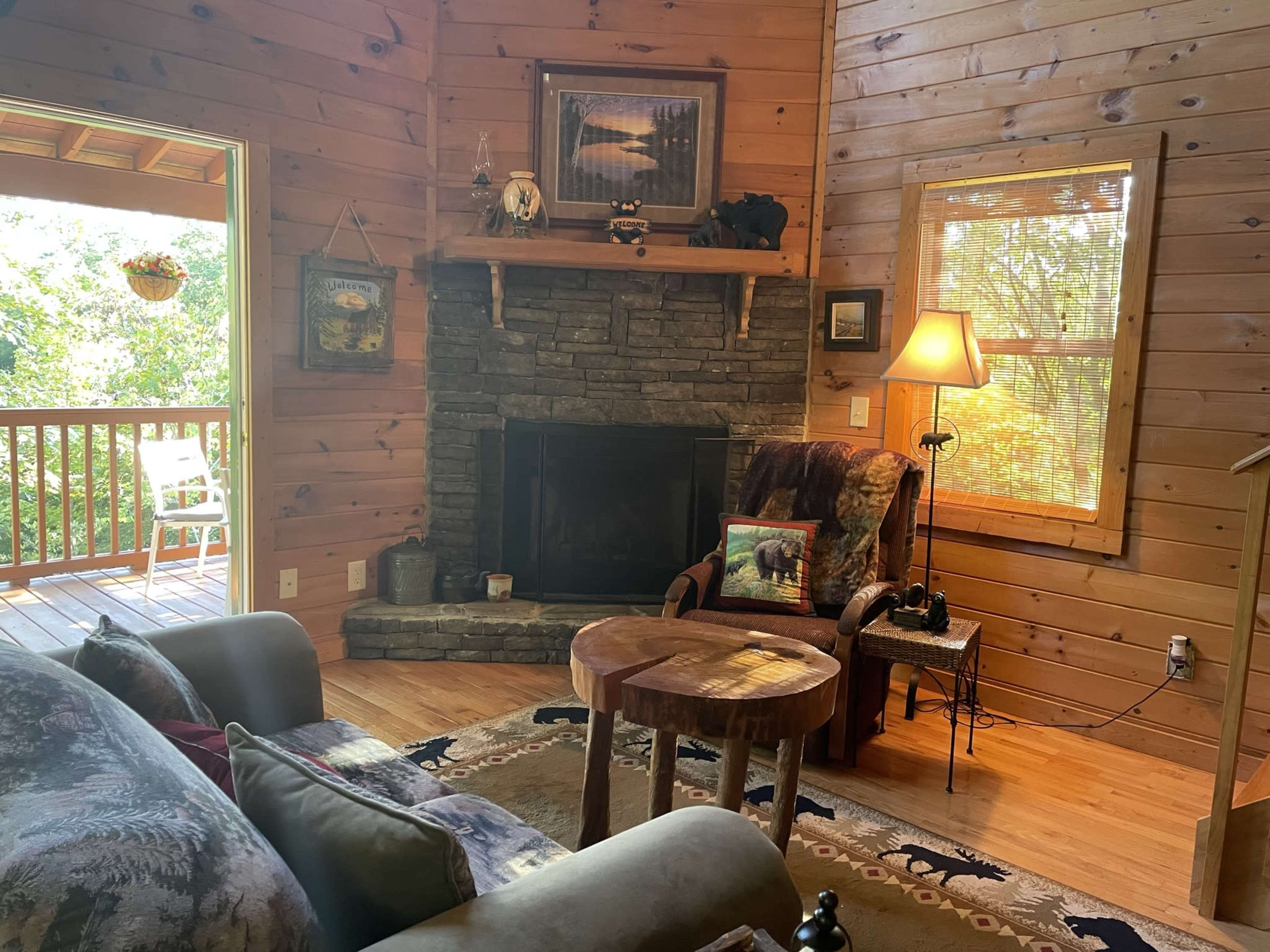 The image shows a cozy wooden living room with a stone fireplace, a comfortable armchair, and large windows overlooking a green outdoor area.