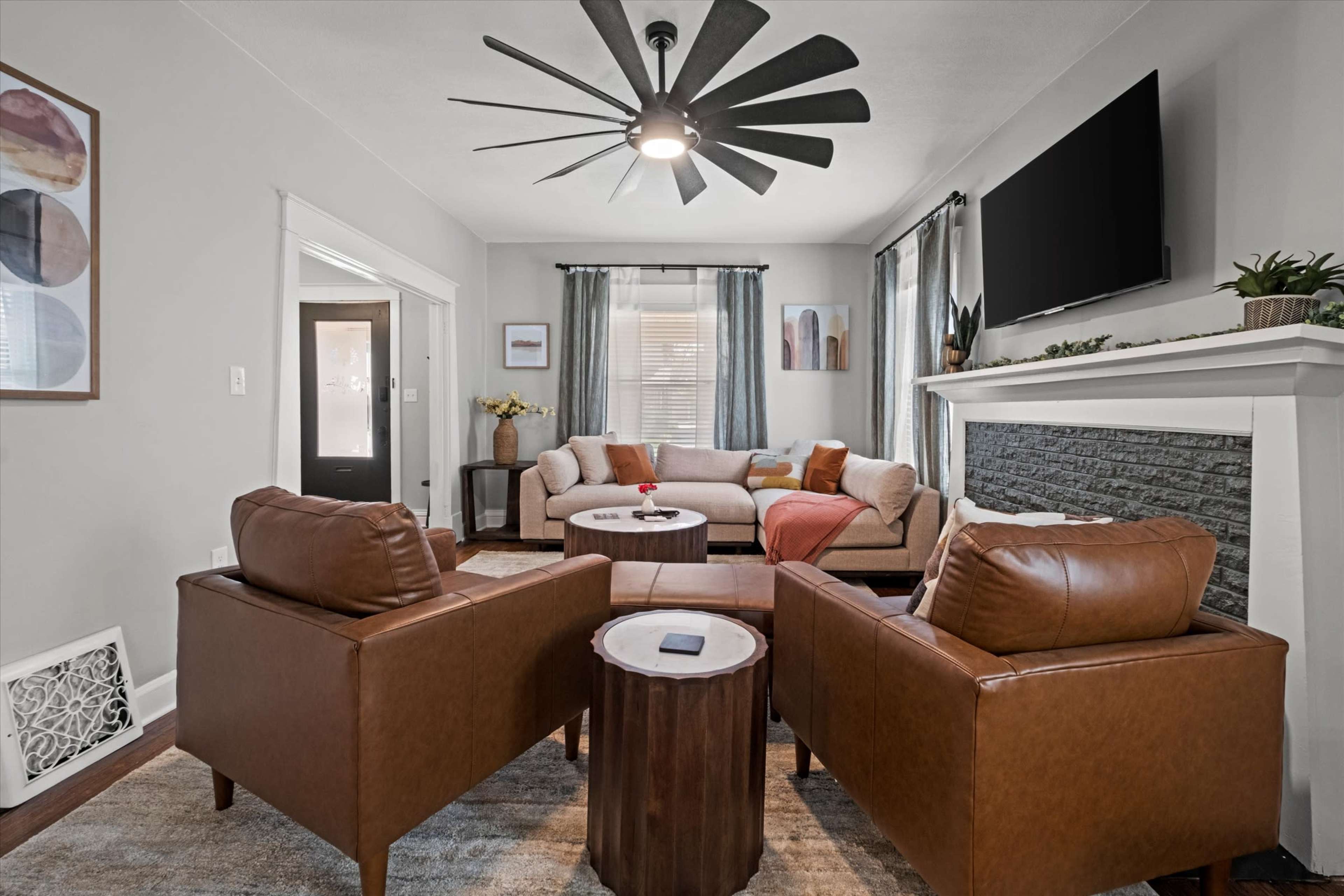The living room features a beige sofa, two brown leather chairs, a round table, and a modern ceiling fan, with art pieces and a television on the walls.