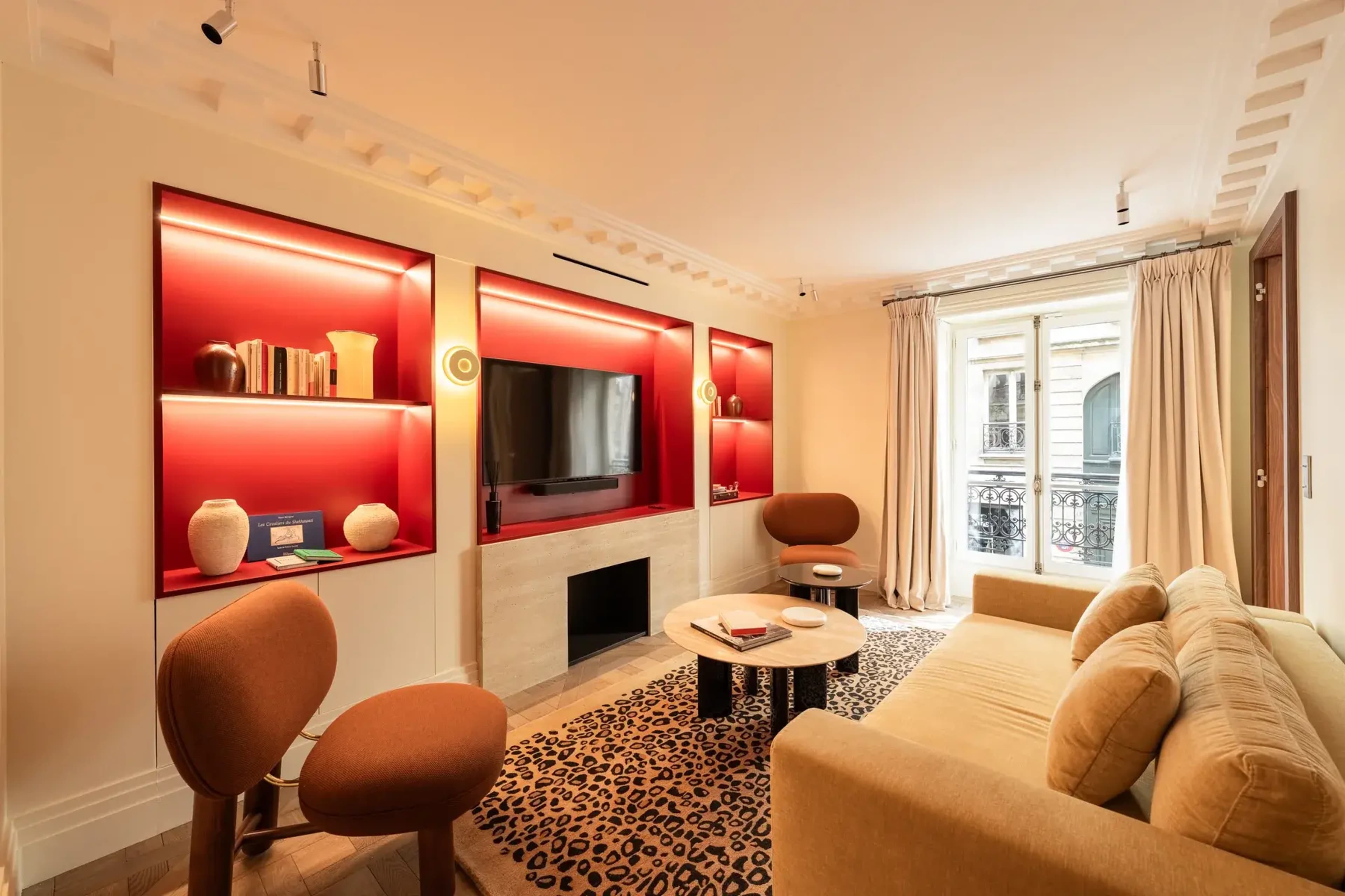 The image shows a modern living room featuring a beige sofa, two brown armchairs, a round coffee table, and a wall-mounted television surrounded by red-backlit shelving.