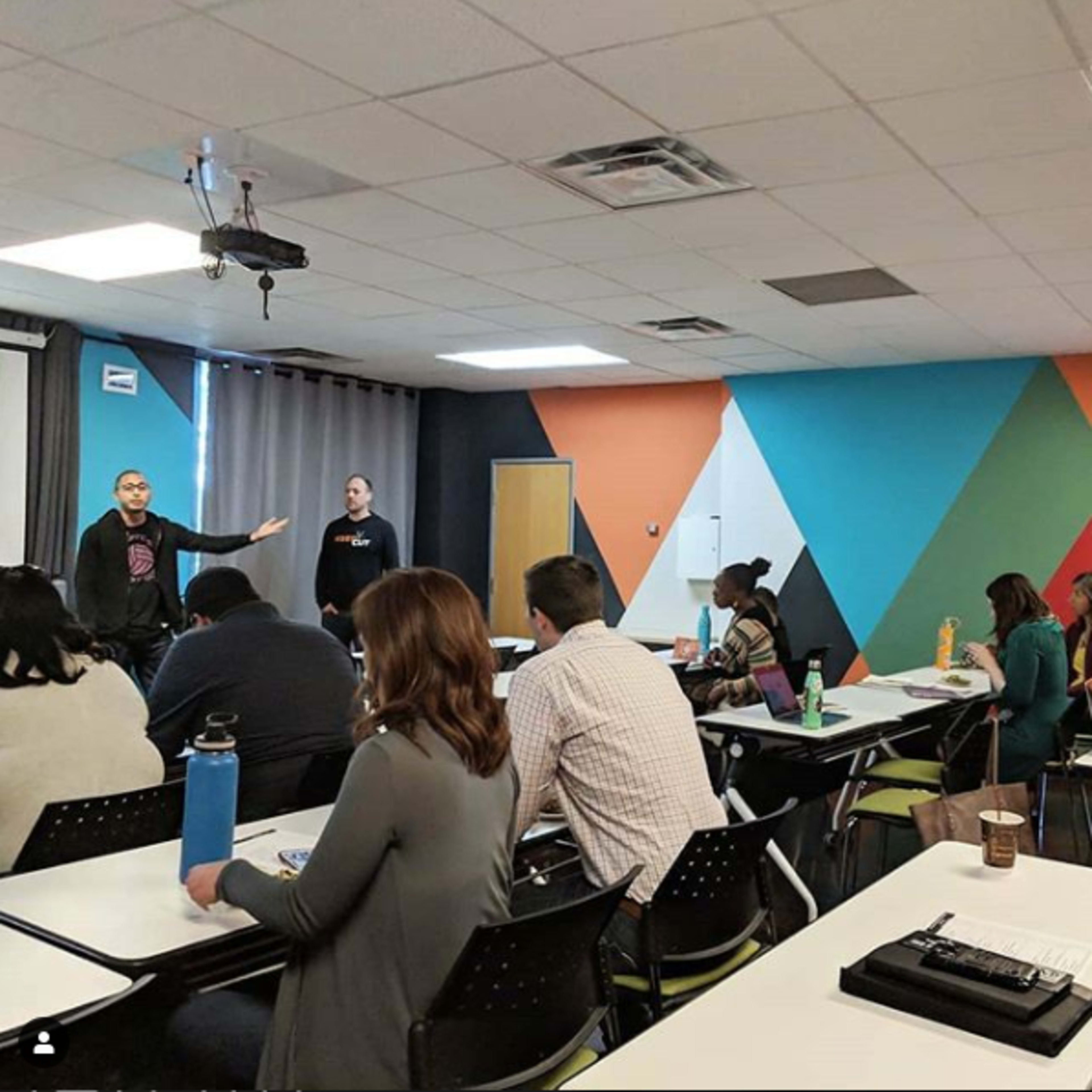 Two speakers are presenting in front of a seated audience in a brightly colored classroom.
