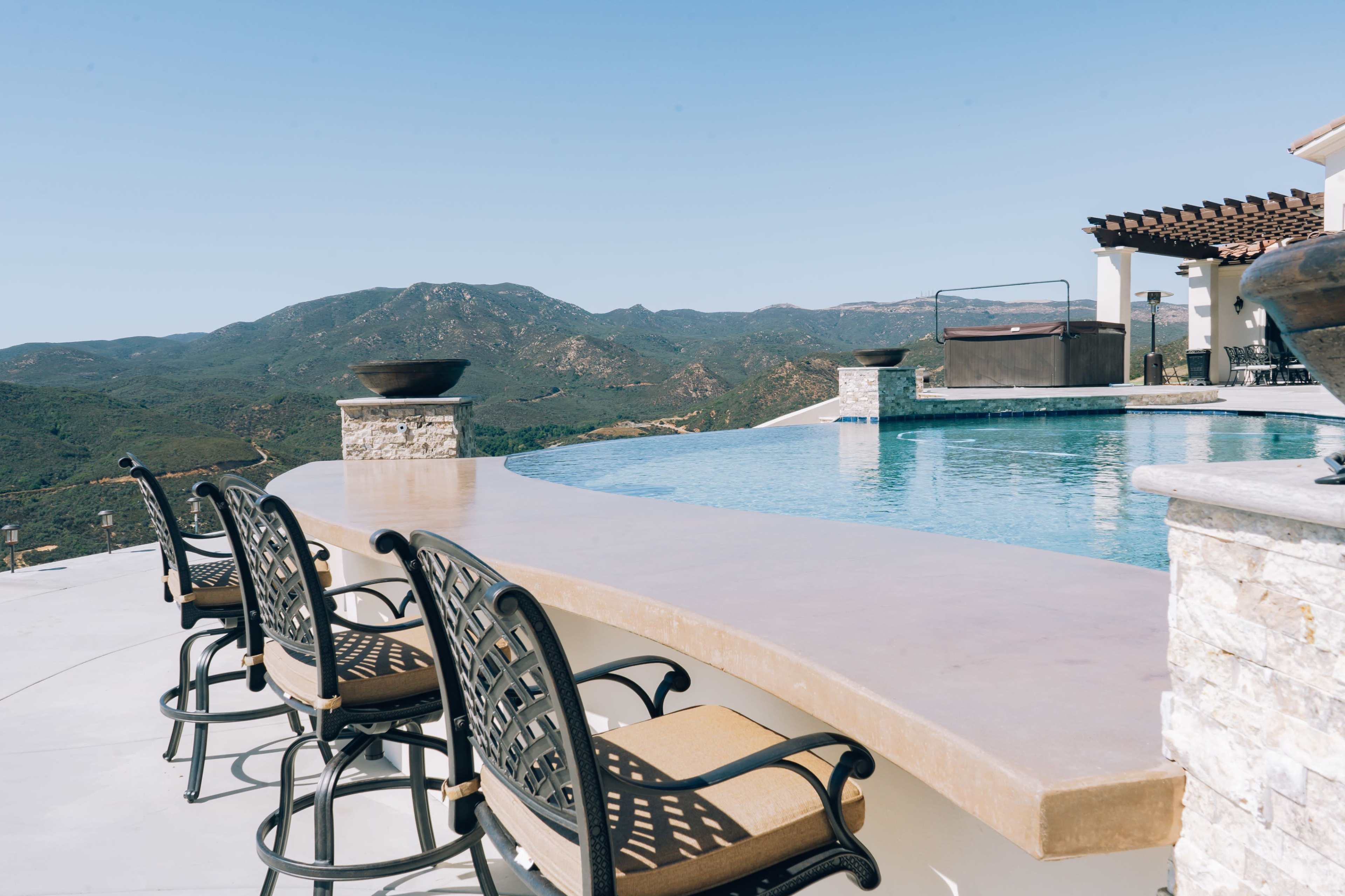 A swimming pool with a curved edge overlooks a mountainous landscape, featuring several barstools along its perimeter.