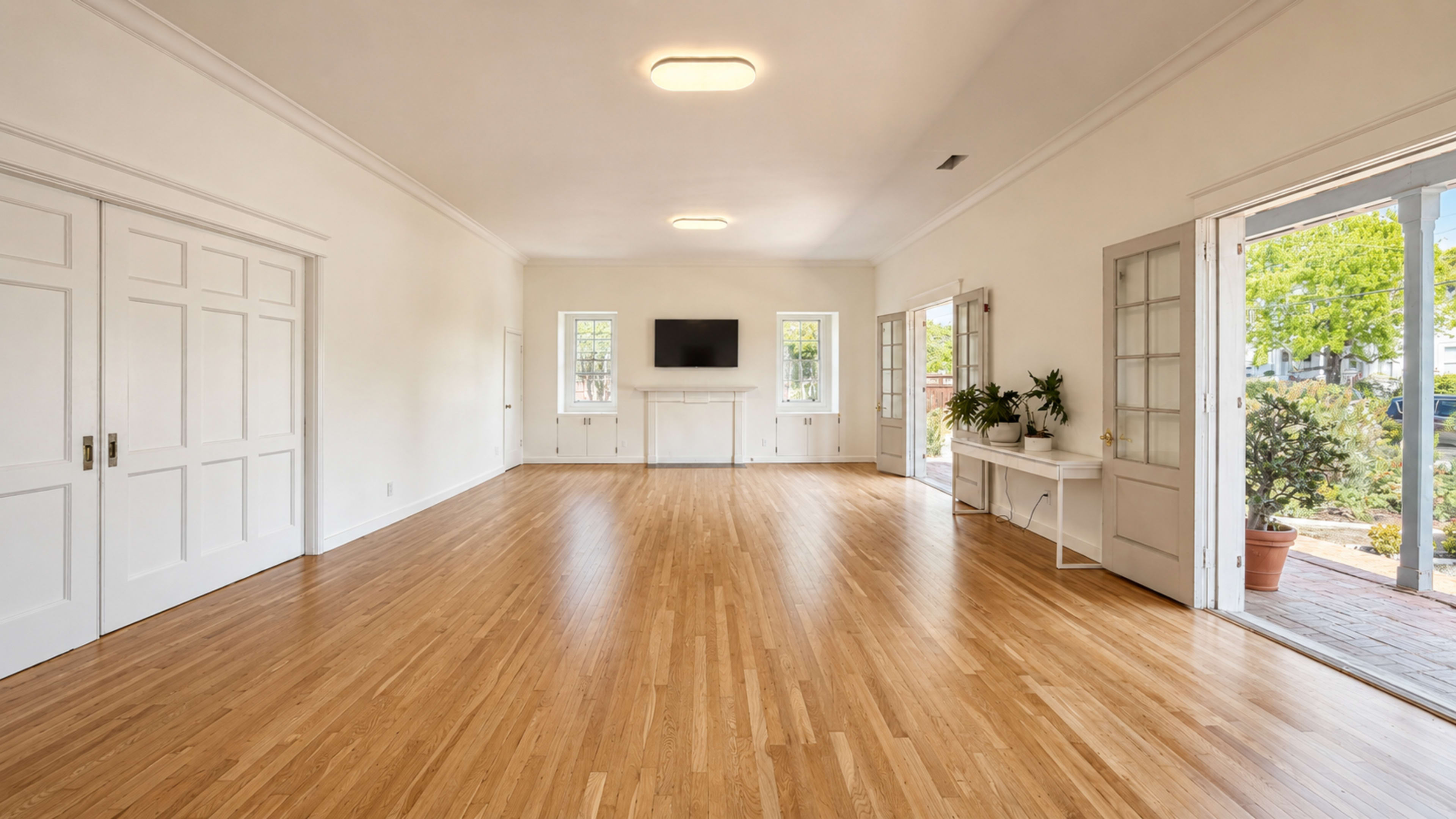 The image shows a spacious, empty room with hardwood flooring, white walls, and large windows allowing natural light, alongside a fireplace and a television mounted on the wall.