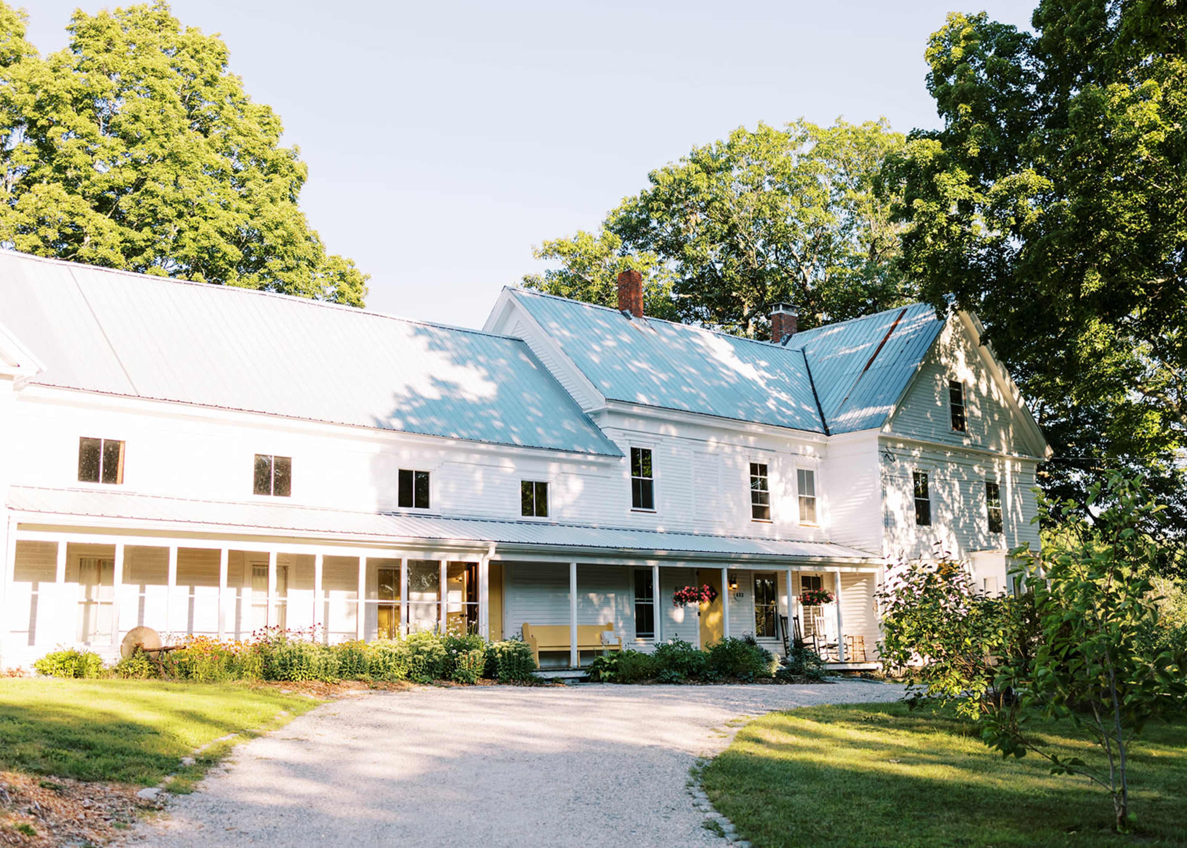 A large white farmhouse with a green metal roof is set amidst trees and a gravel driveway.