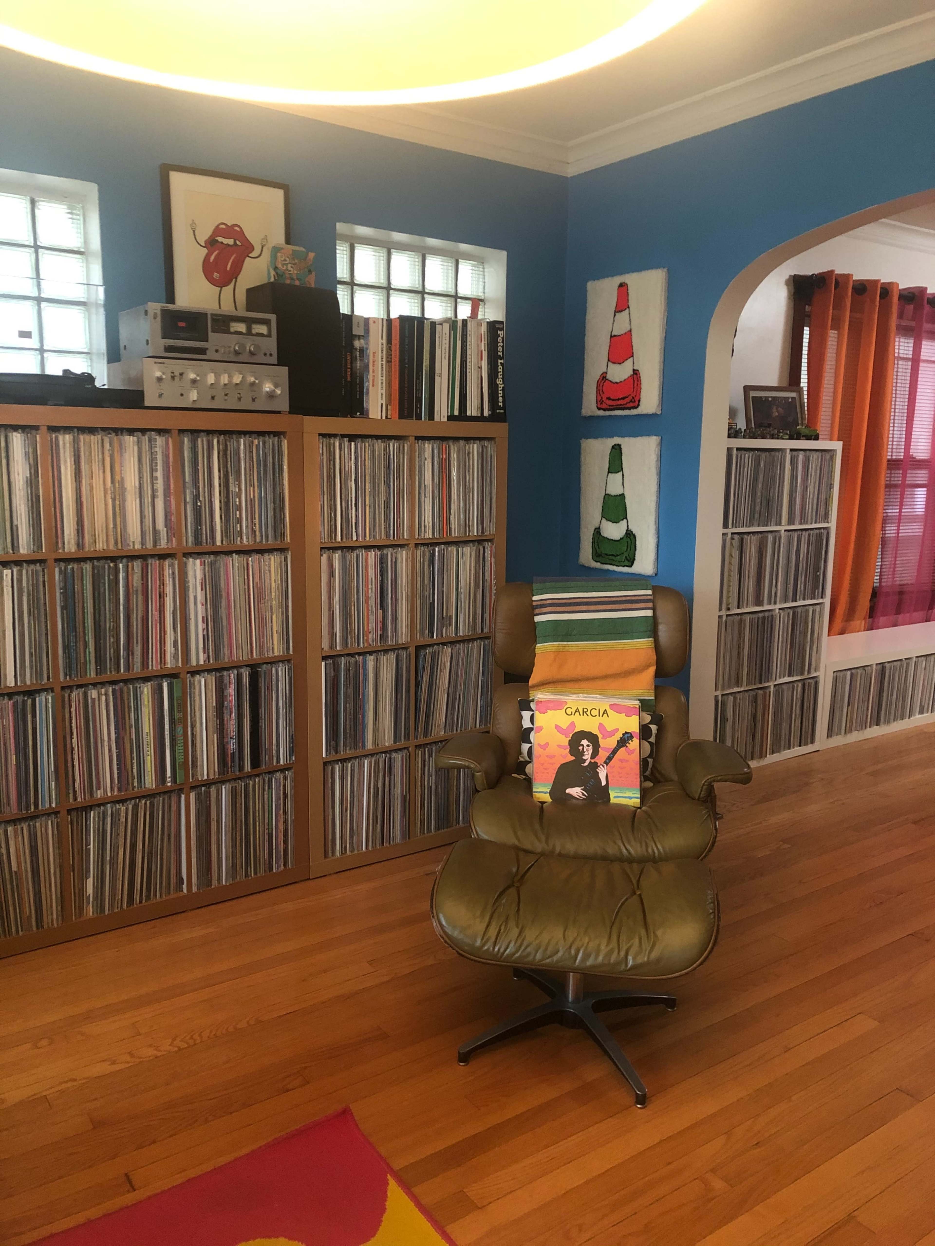 The image shows a room with a green chair positioned in front of wooden shelves filled with vinyl records, alongside colorful paintings and a sound system.