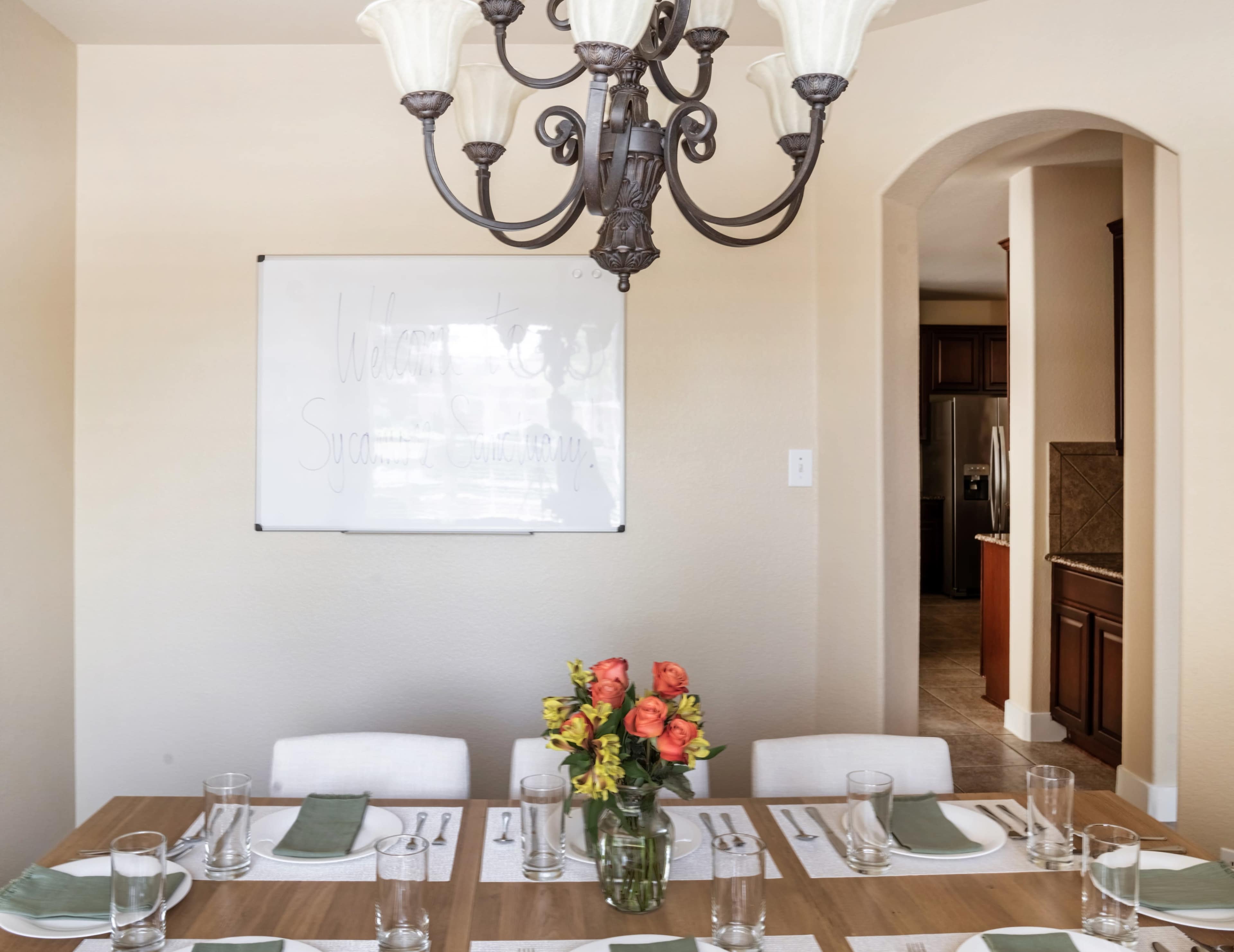 A dining table is set with glassware and linen, featuring a vase of flowers, beneath a chandelier, with a welcome sign on the wall and an adjoining kitchen visible in the background.