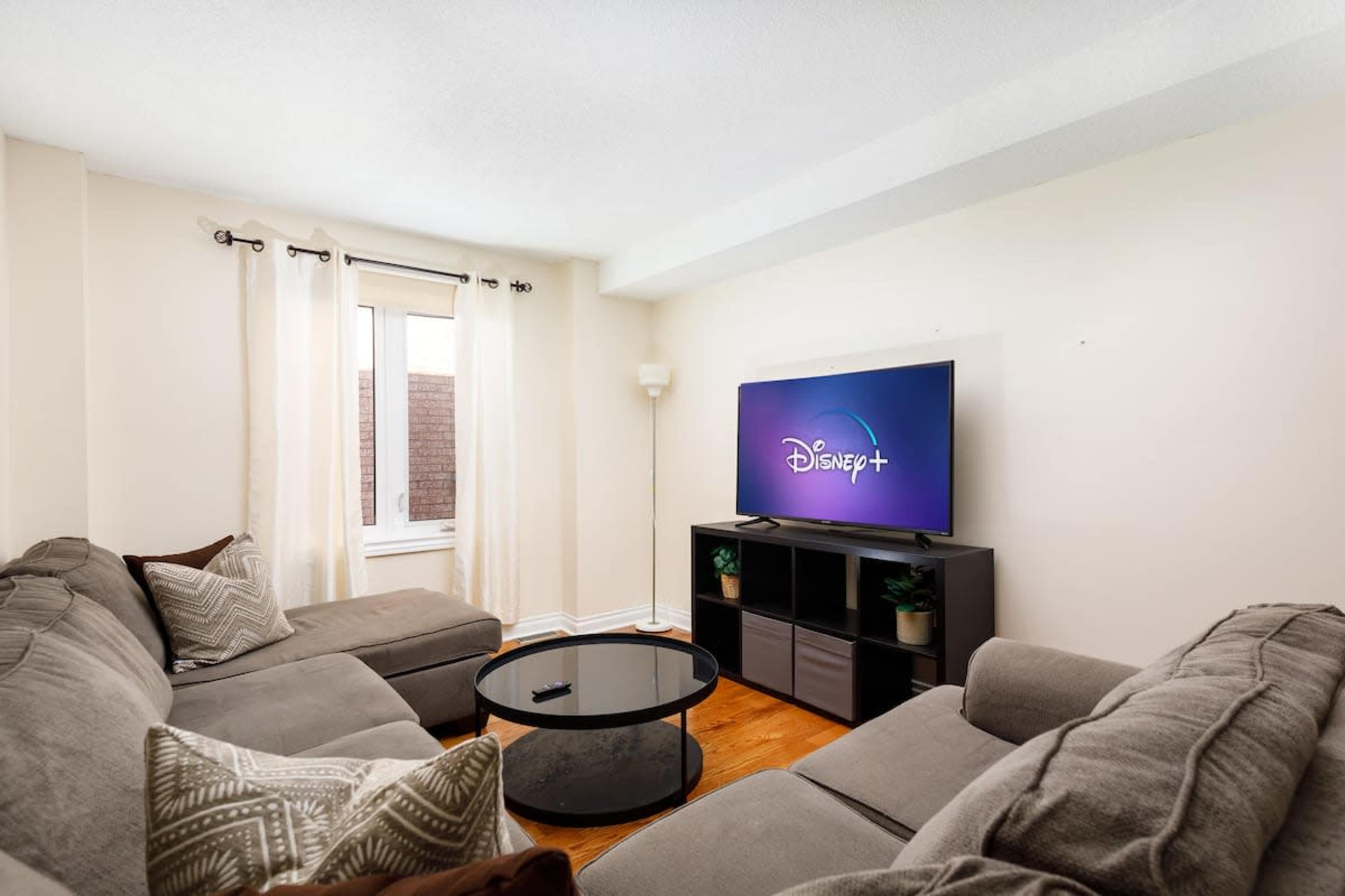 A living room features a gray sectional sofa, a round coffee table, and a television displaying the Disney+ logo, with a window covered by sheer curtains.