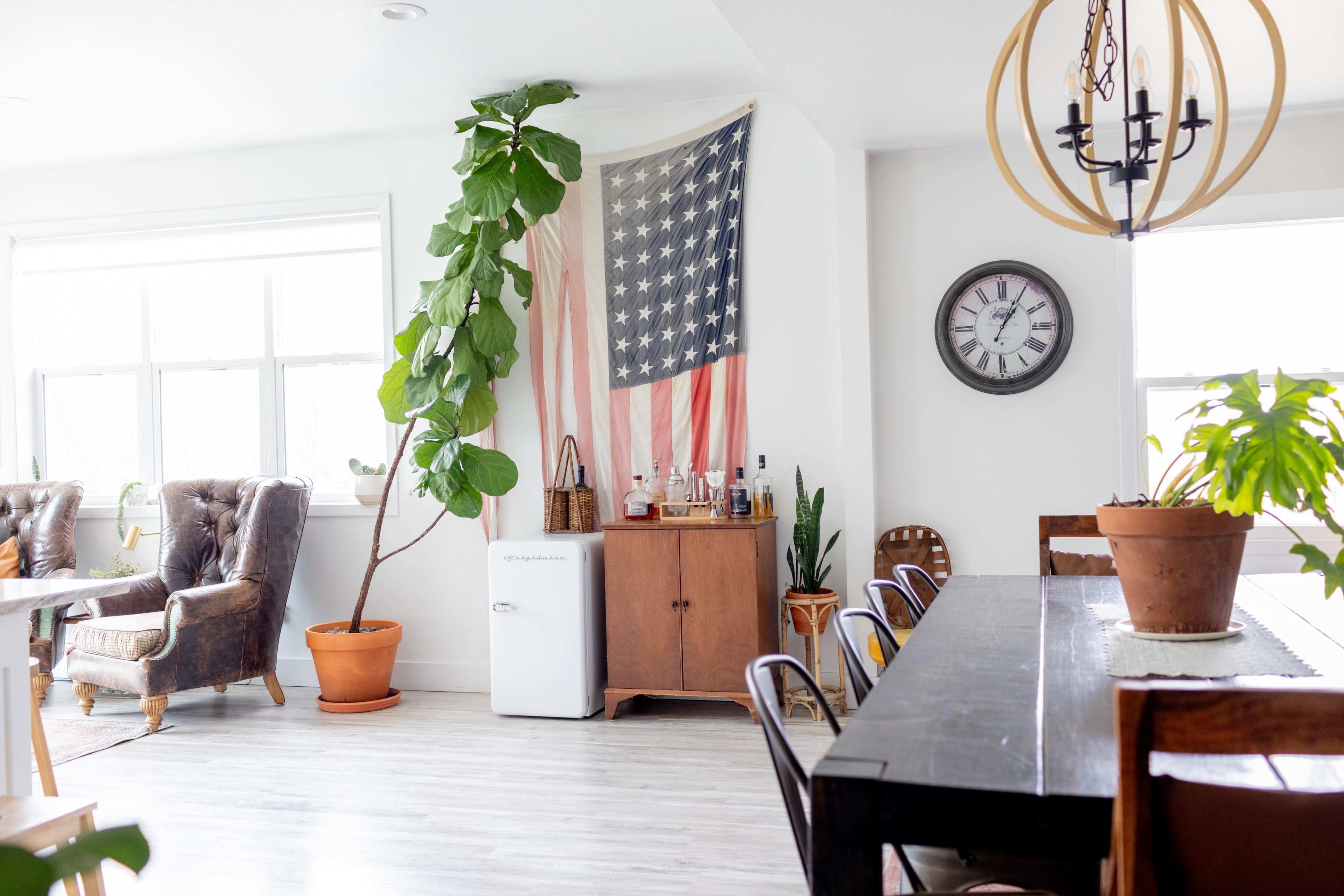 A spacious dining area features a black wooden table, a vintage clock on the wall, a potted plant beside a flag decor, and a small refrigerator near a wooden cabinet.