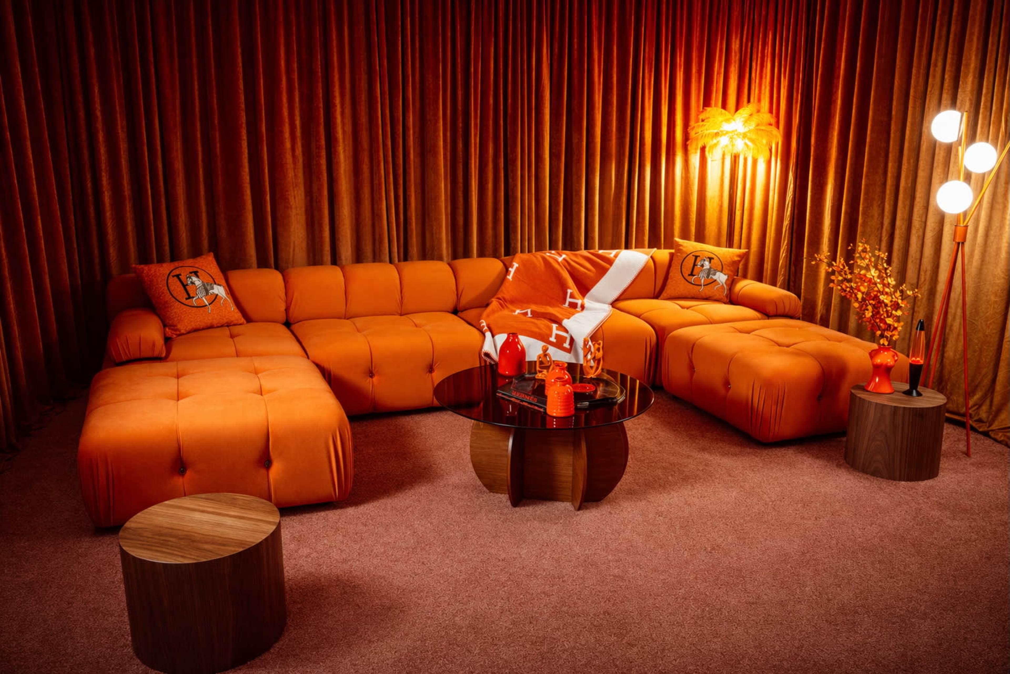 The image features a vibrant orange sectional sofa, accompanied by a round wooden coffee table and illuminated by warm lighting, all set against a backdrop of textured orange curtains.