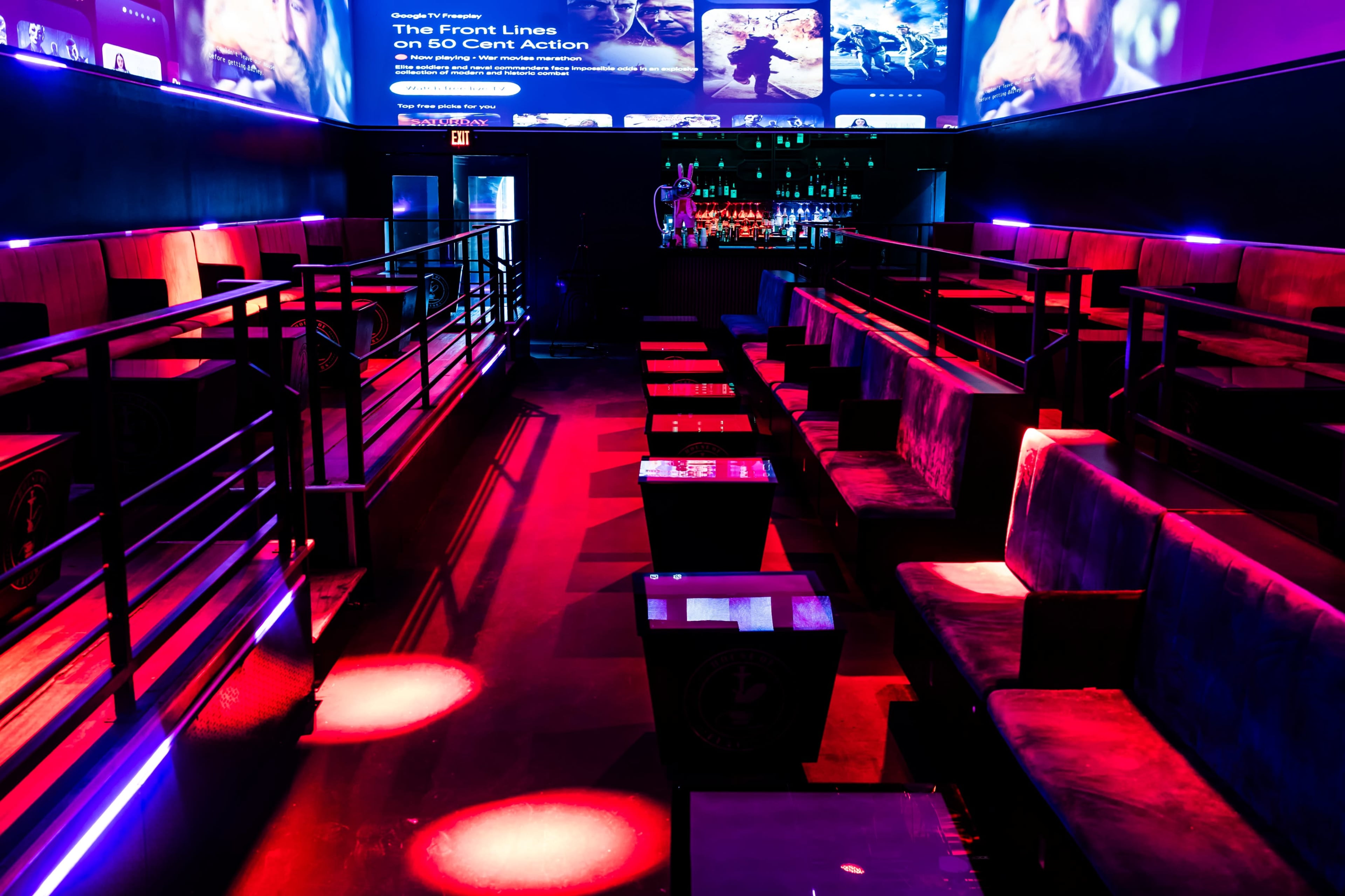 The image shows an empty lounge area with modern seating arrangements, low tables, and colorful lighting effects in a nightlife venue.