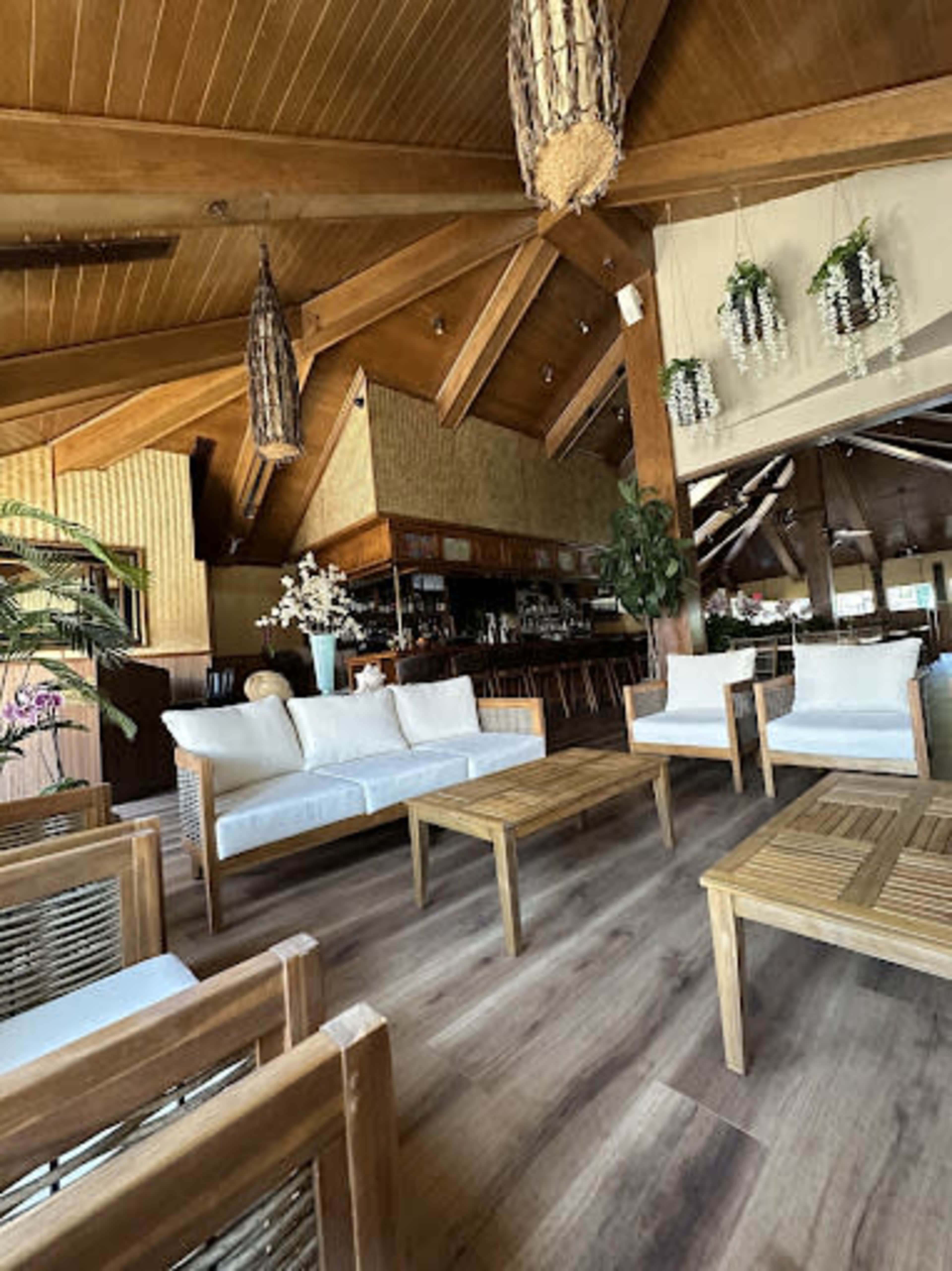 The image shows a cozy seating area with white sofas and wooden tables beneath a wooden ceiling, featuring tropical decor and plants.