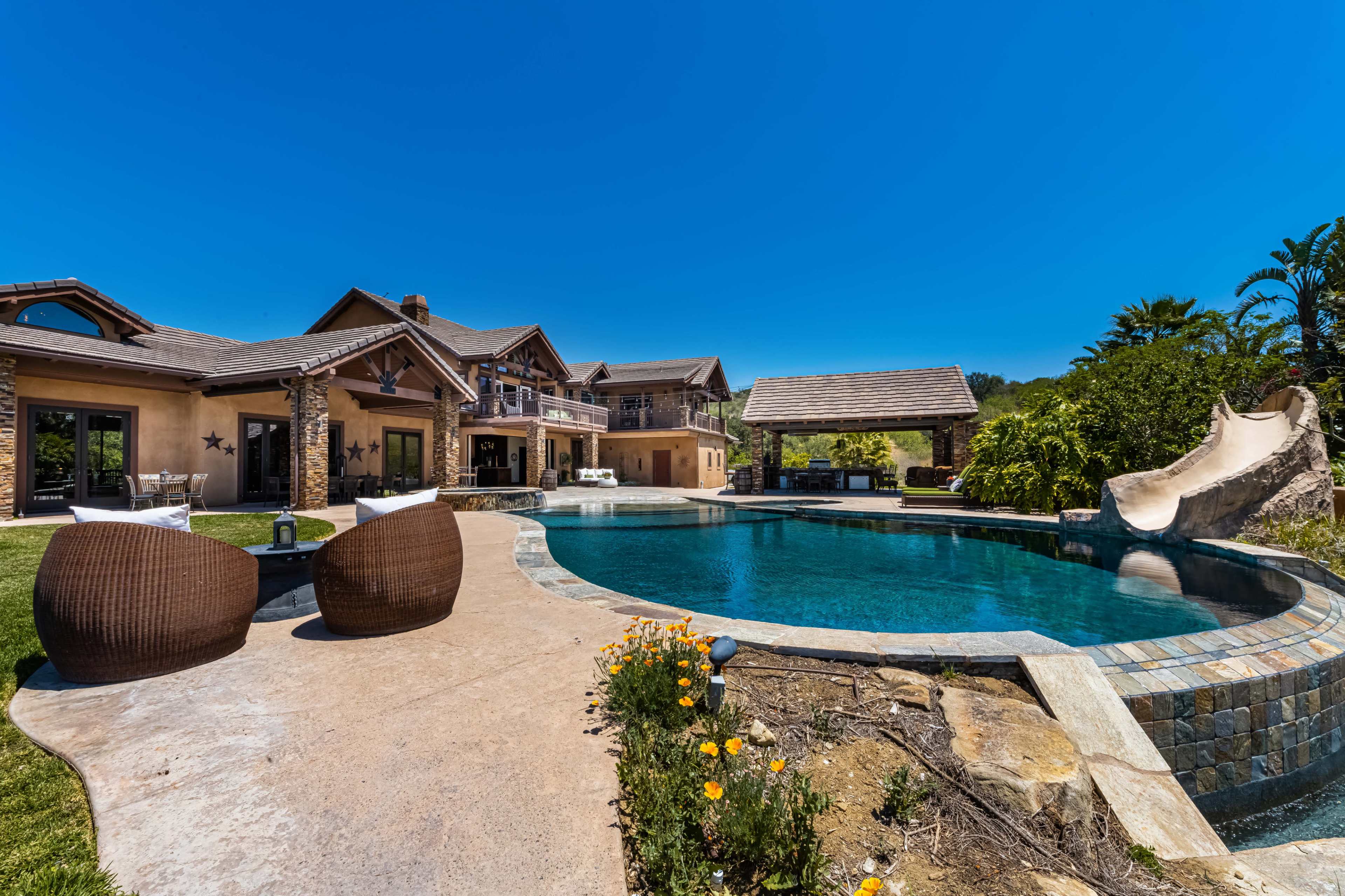 The image shows a spacious backyard featuring a swimming pool with a slide, surrounded by a modern house and lush greenery under a clear blue sky.