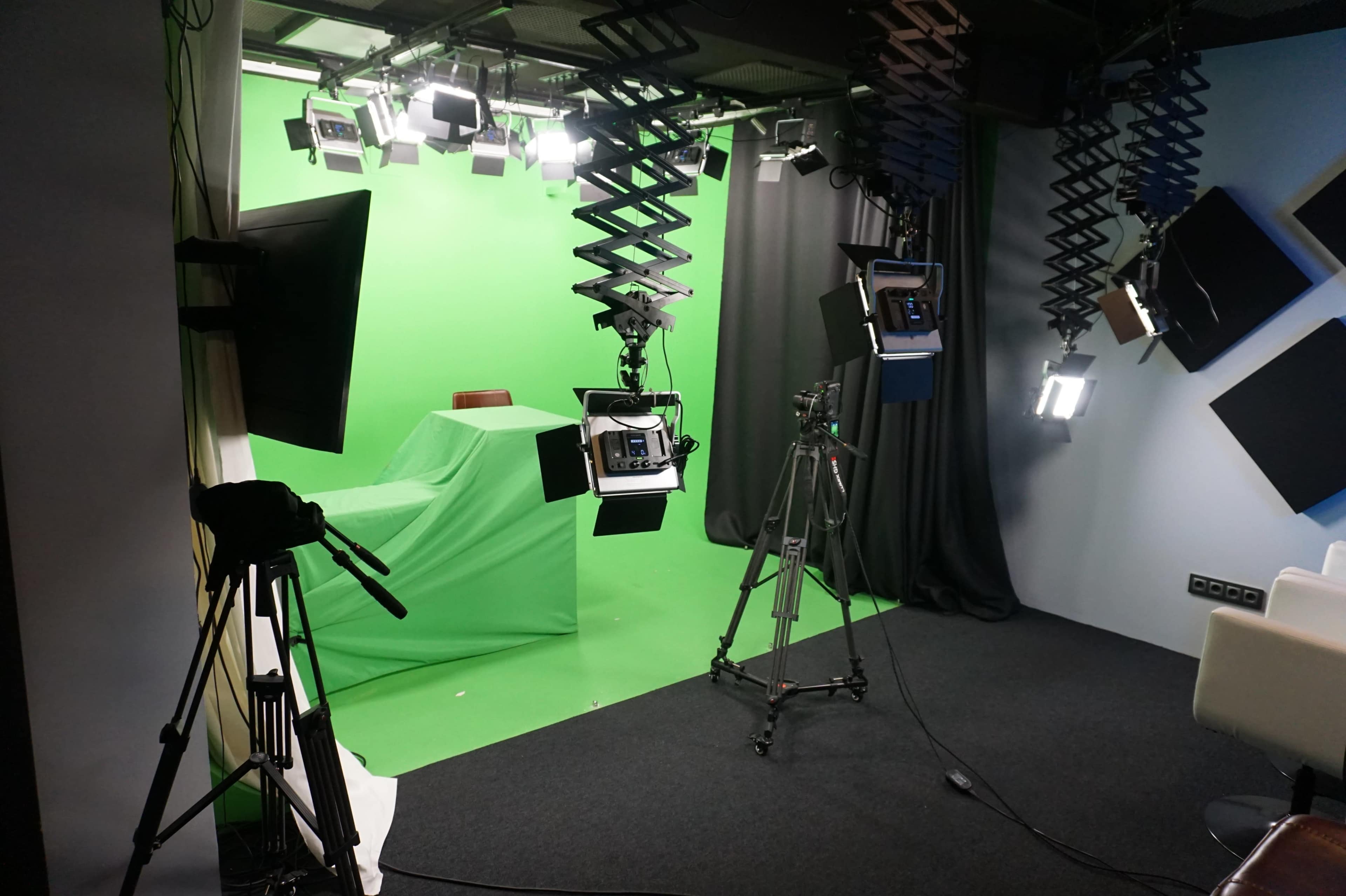 The image shows a studio setup featuring a green screen, professional lighting equipment, and cameras arranged around the space.